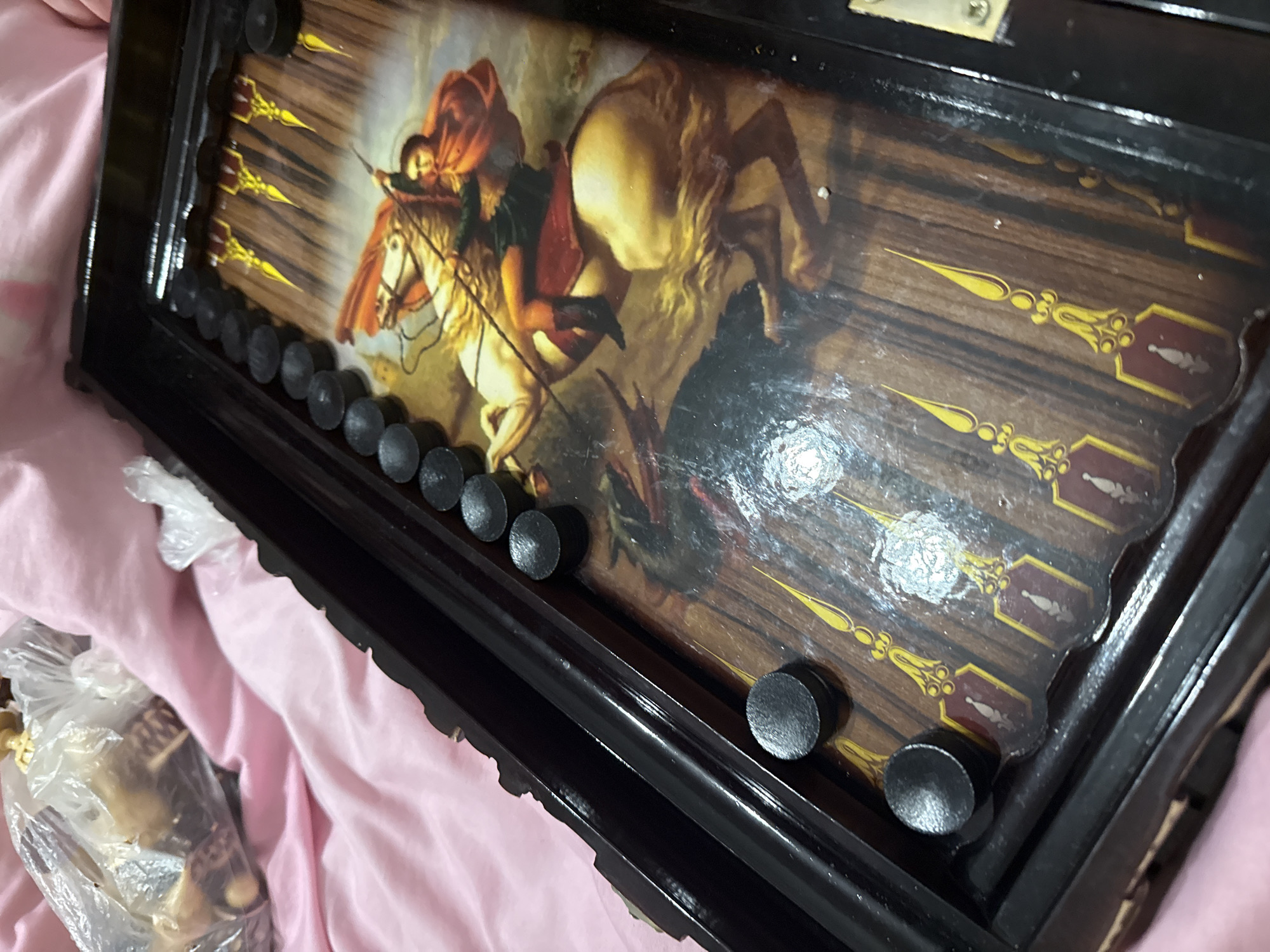 A close-up shot shows a partially opened backgammon board resting on a pink surface. The board is adorned with an image of a knight on horseback slaying a dragon. Black checkers are positioned along the edges of the board. A plastic bag containing indistinguishable items is visible in the lower-left corner of the frame. There is no information in this image to suggest any connection to Khimki, Russia.