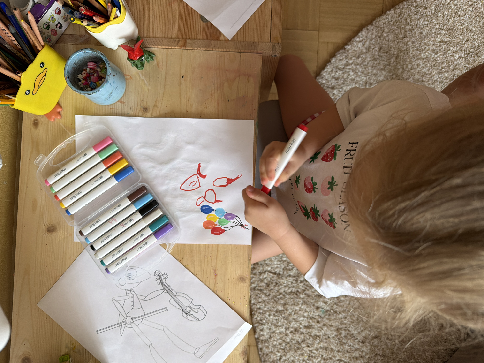 A high-angle shot shows a child coloring at a wooden table, likely in Khimki, Russia, given the "OENMY" marker label. The child, wearing a white shirt with a strawberry pattern, sits on a shaggy rug. On the table are two drawings: one of a figure playing the violin and another of balloons and a heart. Also present are various art supplies, including markers in a plastic case, colored pencils in two pen holders, and a small container filled with objects. The child is using a red marker to color.