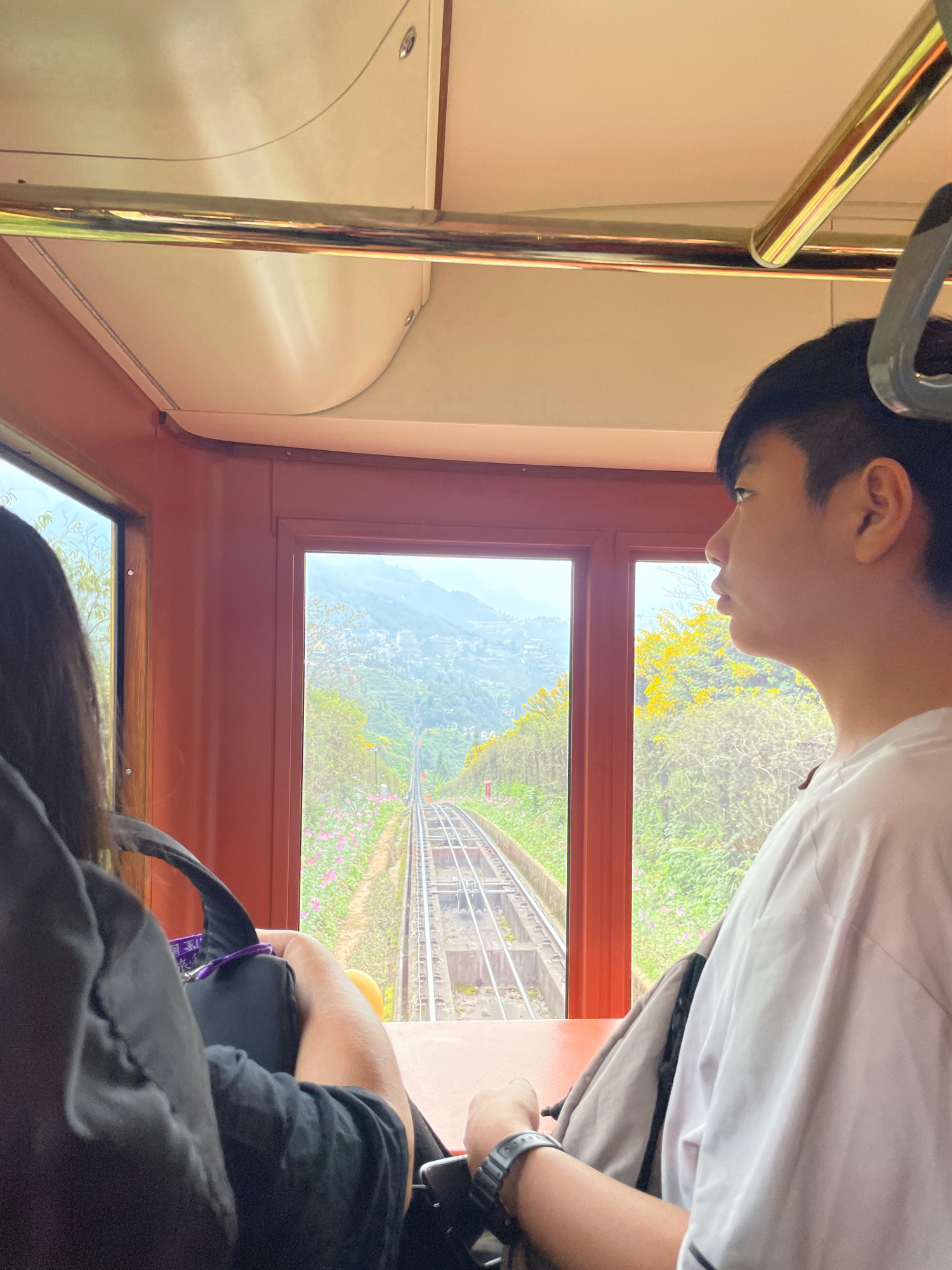 The image depicts the interior of a funicular train ascending a steep mountain in Sa Pa Ward, Vietnam. Two individuals are seated inside. On the left, a person with long dark hair, wearing a dark top, is partially visible, holding a black bag with a purple strap. On the right, a young person with short dark hair, wearing a white t-shirt, gazes out the window. The funicular's windows offer a view of the tracks descending into a valley. The landscape outside is characterized by terraced hillsides dotted with buildings, lush green vegetation, and blooming yellow and pink flowers. The interior of the funicular features light-colored upholstery and polished metal handrails. The perspective suggests the photo was taken from within the train, looking forward and slightly to the right.