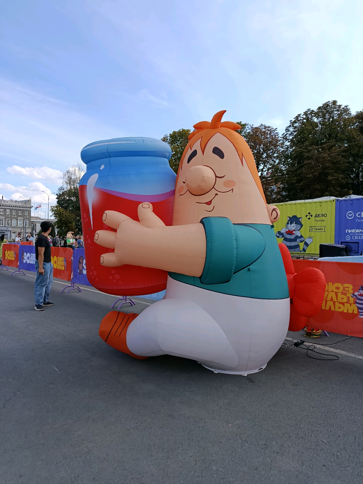 An inflatable cartoon character, resembling a young boy with orange hair and a rosy complexion, is depicted holding a giant red jar filled with a red liquid. The character is seated on the ground. The scene appears to be outdoors, with a paved surface in the foreground and trees and buildings visible in the background. There are also several banners and what seem to be event barriers in the middle ground. A person in a dark t-shirt and jeans stands to the left of the inflatable, looking towards it. The event appears to be taking place in the city of Saratov, Russia, as indicated by the presence of a banner with Cyrillic text and a recognizable building in the background consistent with Russian architecture. The sky is partly cloudy.