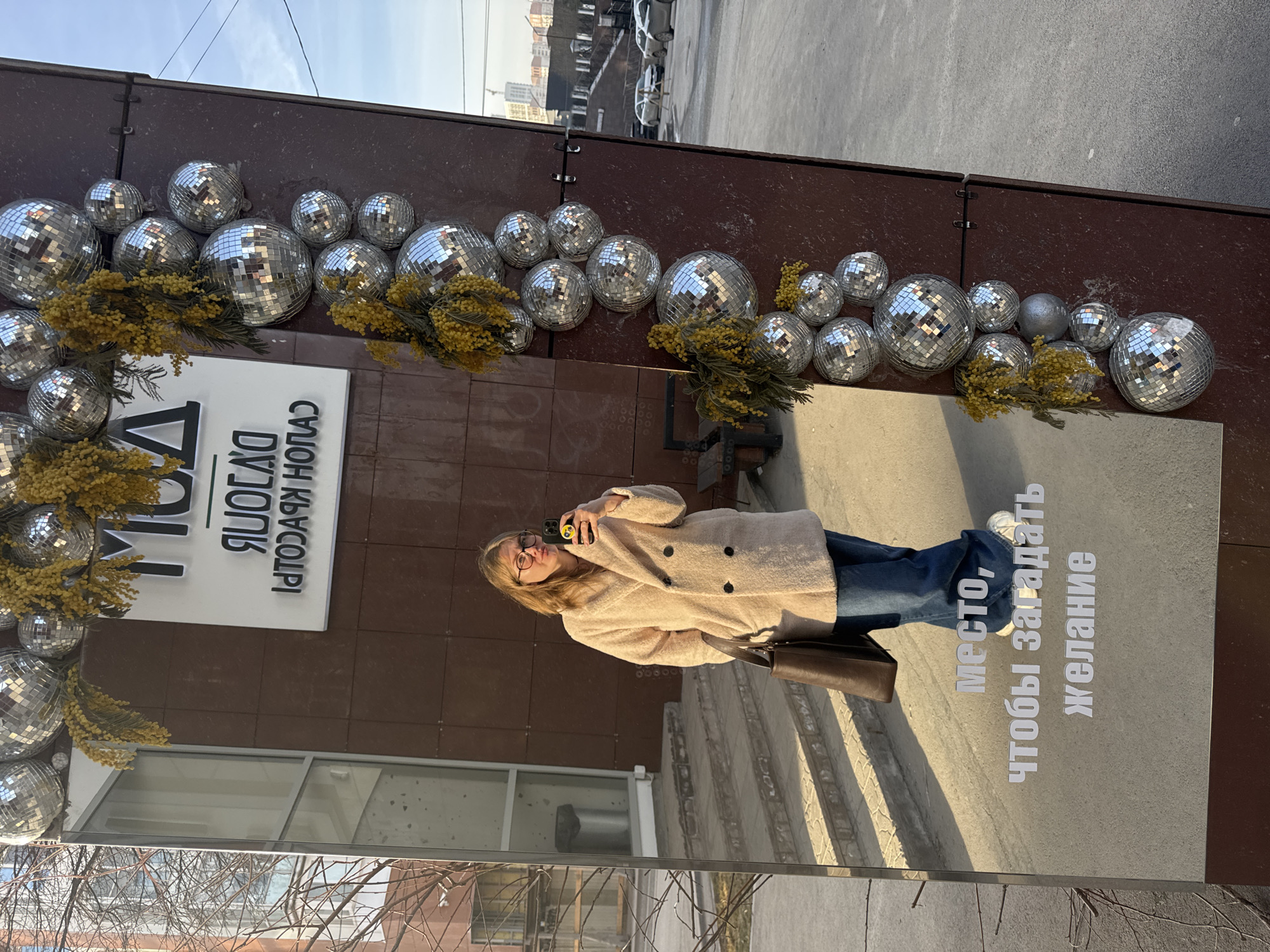 A woman is standing in front of a building with disco ball decorations in Perm, Russia. She is taking a picture of herself in a mirrored panel that is attached to the building. The sign on the mirrored panel reads, "Place to make a wish." The building has a sign in Russian above the mirrored panel that says "DA DNYA KRASOTY SALON." The woman is wearing a light-colored coat, blue jeans, and white sneakers. She has a brown bag over her shoulder. The street behind the building has cars and buildings. The sky is clear.