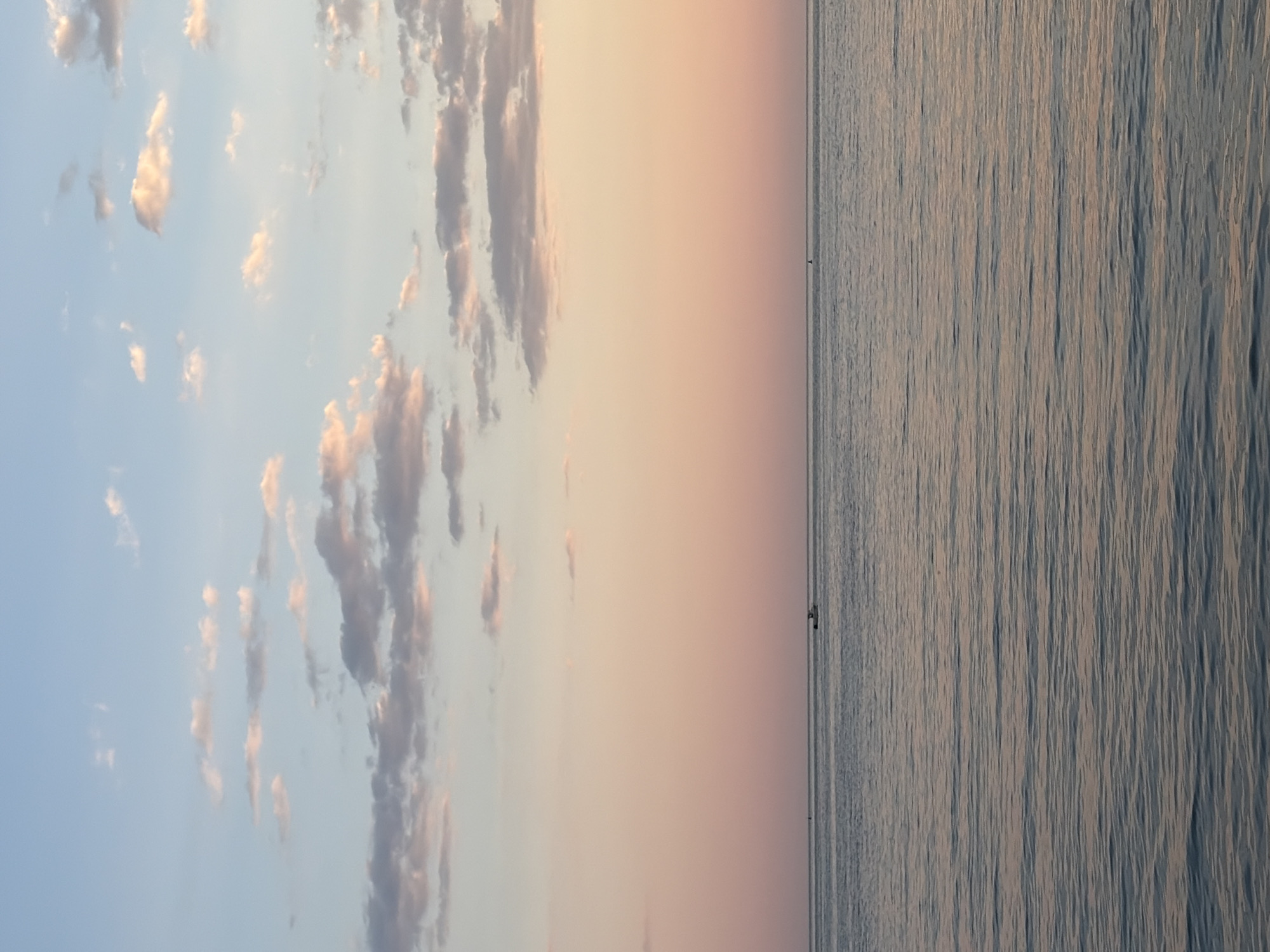 The image is a landscape of a sunrise or sunset over the sea, possibly in Budva, Montenegro. The left side of the image features a light blue sky with scattered white and gray clouds, tinged with pink from the sun. The middle section shows the sky transitioning into a soft gradient of pink and orange hues. The right side depicts the sea, with horizontal lines indicating gentle waves or ripples. There are what appear to be small boats or objects in the distance on the water. The image is oriented vertically.