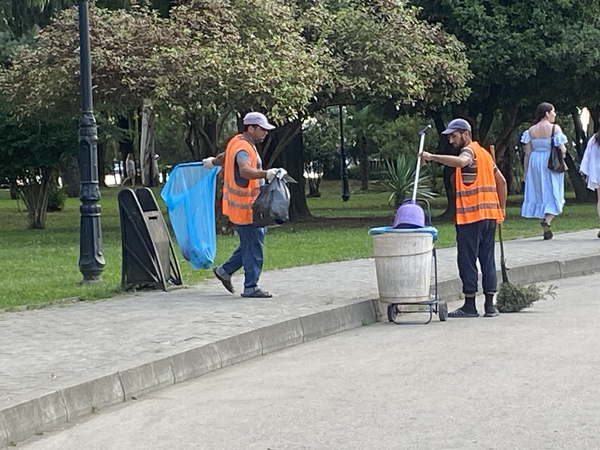 In Sokhumi, Abkhazia, two individuals wearing orange safety vests are engaged in cleaning a paved walkway in a park-like setting. The person on the left is holding a large blue plastic bag and a black trash bag, with a trash receptacle visible behind them. The person on the right is holding a broom and standing next to a wheeled trash bin. In the background, a woman in a light blue dress walks away from the camera, and other people are present further in the background. The environment includes lush green grass, trees, and a lamppost.