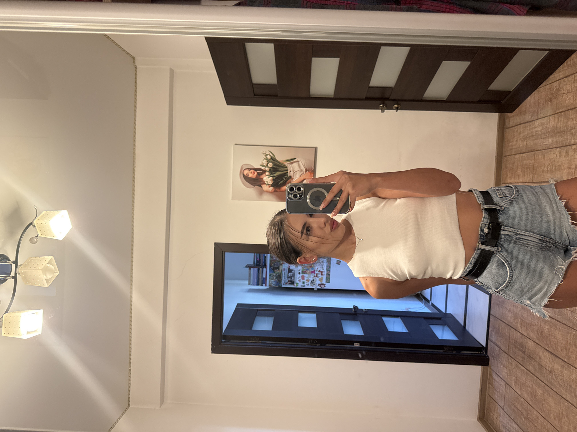 The image is a full-body mirror selfie taken indoors, likely in a home hallway or entryway. A young woman with her hair pulled back is wearing a white crop top and denim shorts with a black belt. She is holding a smartphone to take the photo. In the background, there's an open door revealing a room with shelves filled with books and other items. Another closed door with glass inserts is visible to the right. A framed picture of a woman holding tulips hangs on the wall. The flooring is made of wooden planks. The lighting appears to be from a ceiling fixture with three lamps. The scene is consistent with an interior residential setting.