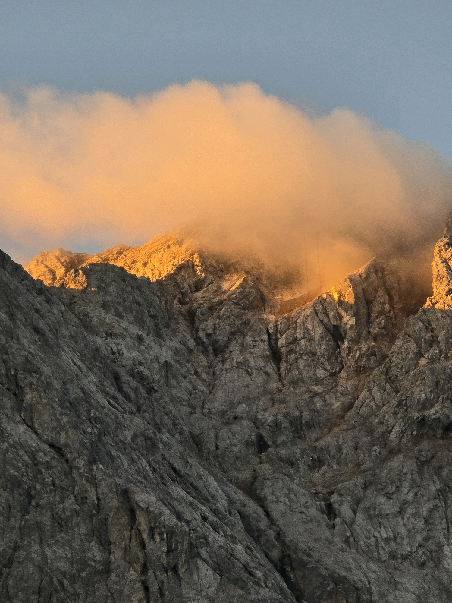 The image shows a close-up view of a rocky mountain face in Grainau, Germany, bathed in the warm, golden light of sunset. The foreground is dominated by the rugged texture of the mountain's grey rock, with deep crevices and sharp edges. Above the mountain peaks, a thick band of clouds is illuminated by the setting sun, casting an orange glow that transitions to yellow and white. The sky above the clouds is a clear, pale blue. There are no people or man-made objects visible in the image.
