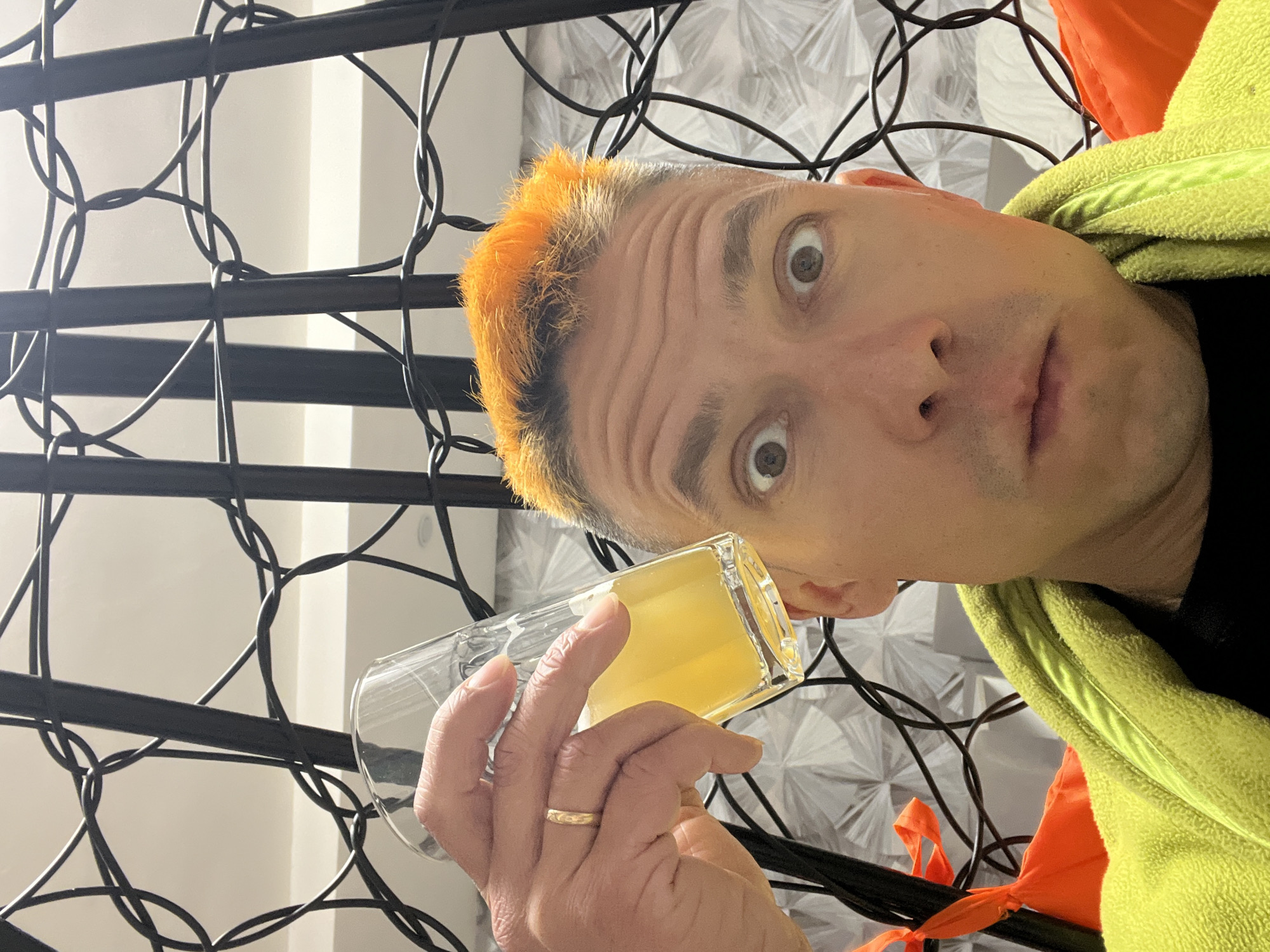 A man with orange hair and wide eyes is holding a glass of yellow liquid near his face. He is wearing a lime green jacket over a black shirt. The background features a black metal trellis with decorative loops, possibly a wine rack or room divider, and a white wall with a geometric pattern. There is an orange tie or ribbon partially visible. The location, Chaikovsky, Russia, cannot be directly determined from this single image.