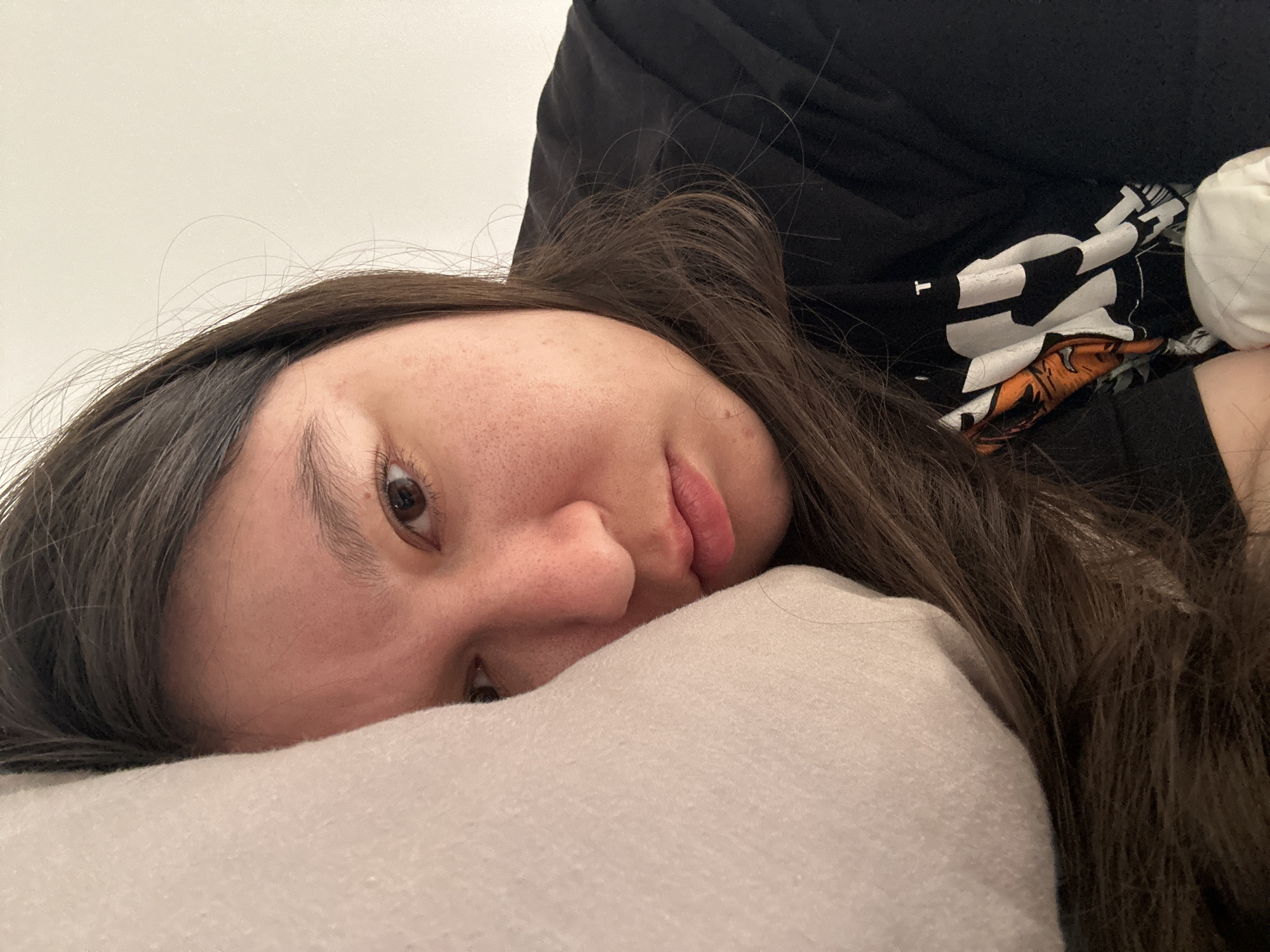 A woman with long, brown hair is lying on a light-colored pillow. The woman's face is turned towards the camera, revealing her nose, lips, and one visible eye. She is wearing a black shirt with white and orange graphics on it. The background is a plain white wall. The image was not taken in Câmara de Lobos, Portugal.