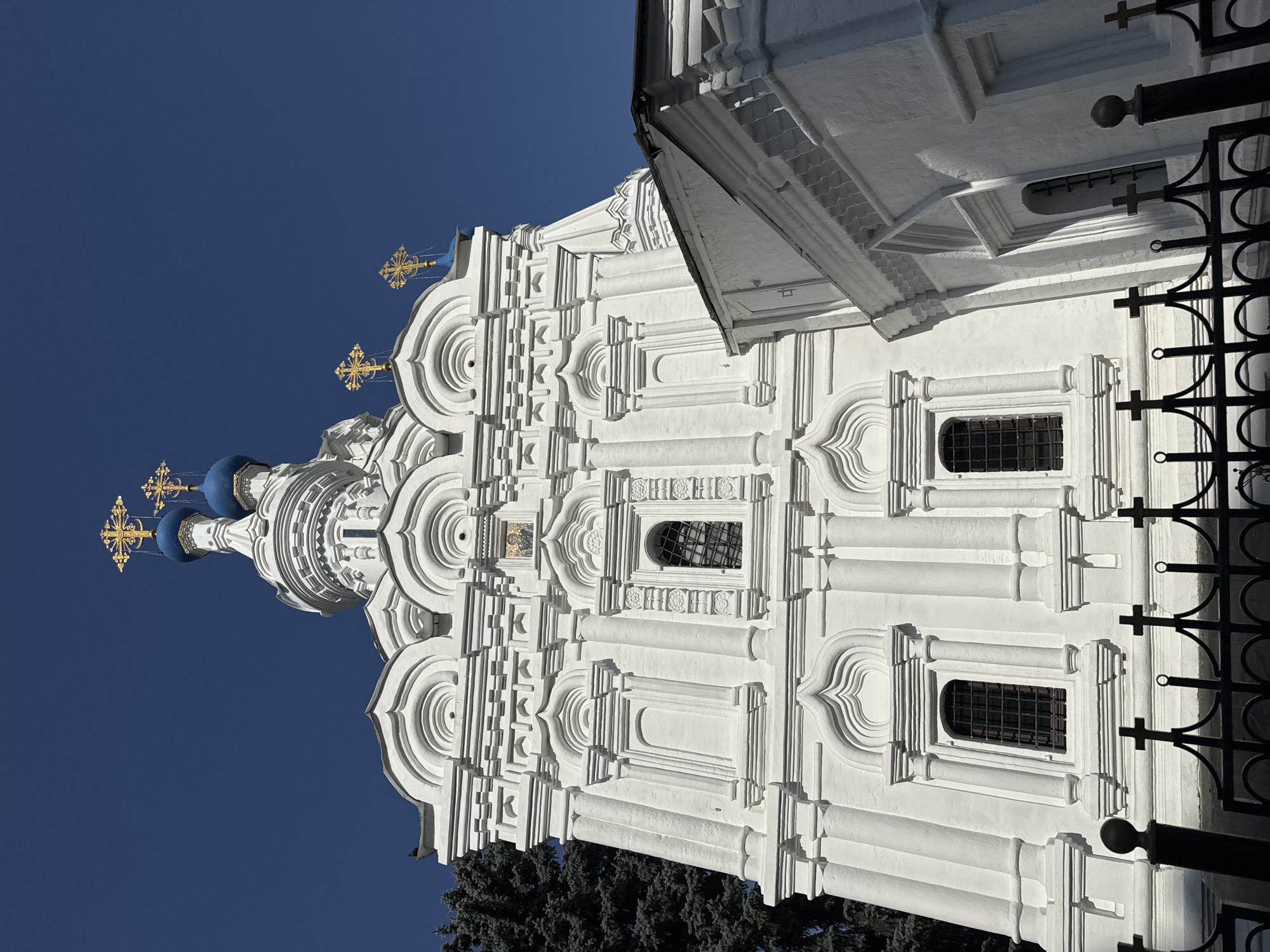 The photograph captures a detailed, low-angle view of the Church of the Dormition of the Mother of God in Kolomenskoye, Moscow, Russia. The white stone church features intricate architectural details, including decorative cornices, recessed niches, and arched window frames. Two prominent blue domes topped with golden crosses are visible against a clear, deep blue sky. A dark green coniferous tree stands to the left of the church, partially obscuring the building. In the foreground, a black metal fence with cross-shaped finials runs horizontally across the bottom of the frame, casting shadows on the ground. The overall scene is bright and sunny, with strong shadows indicating the time of day. There are no people visible in the image.