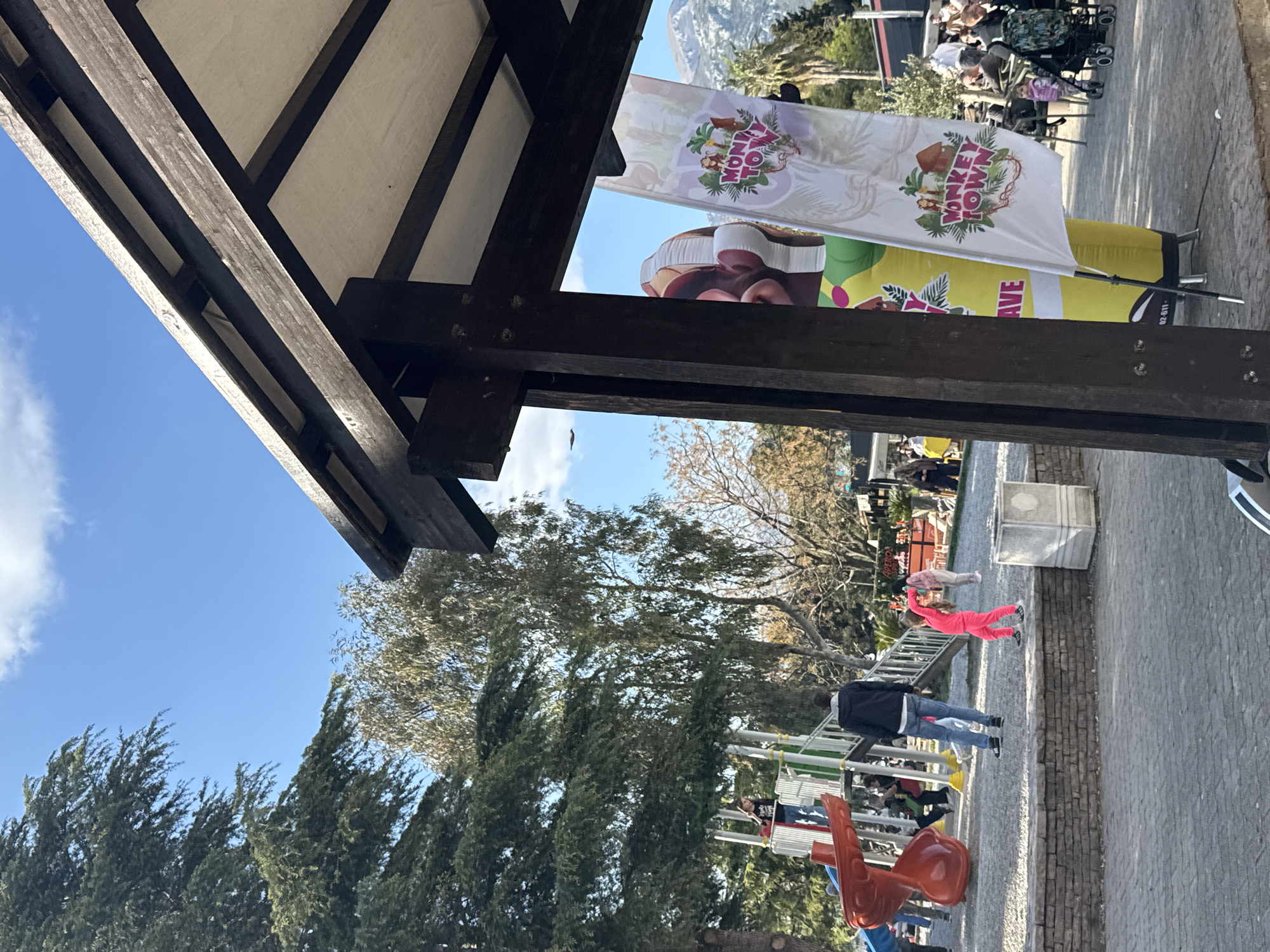 The image, taken in Bar, Montenegro, presents a daytime outdoor scene. A dark wooden structure frames the left portion, while a partially visible "Monkey Town" banner stands in the midground. A playground is visible in the midground with children playing. A person in a pink jumpsuit is walking on the ground. The ground is paved with rectangular stones. Trees and a partial view of a mountain exist in the background. A few adults appear to be supervising children. The sky is blue with scattered clouds.