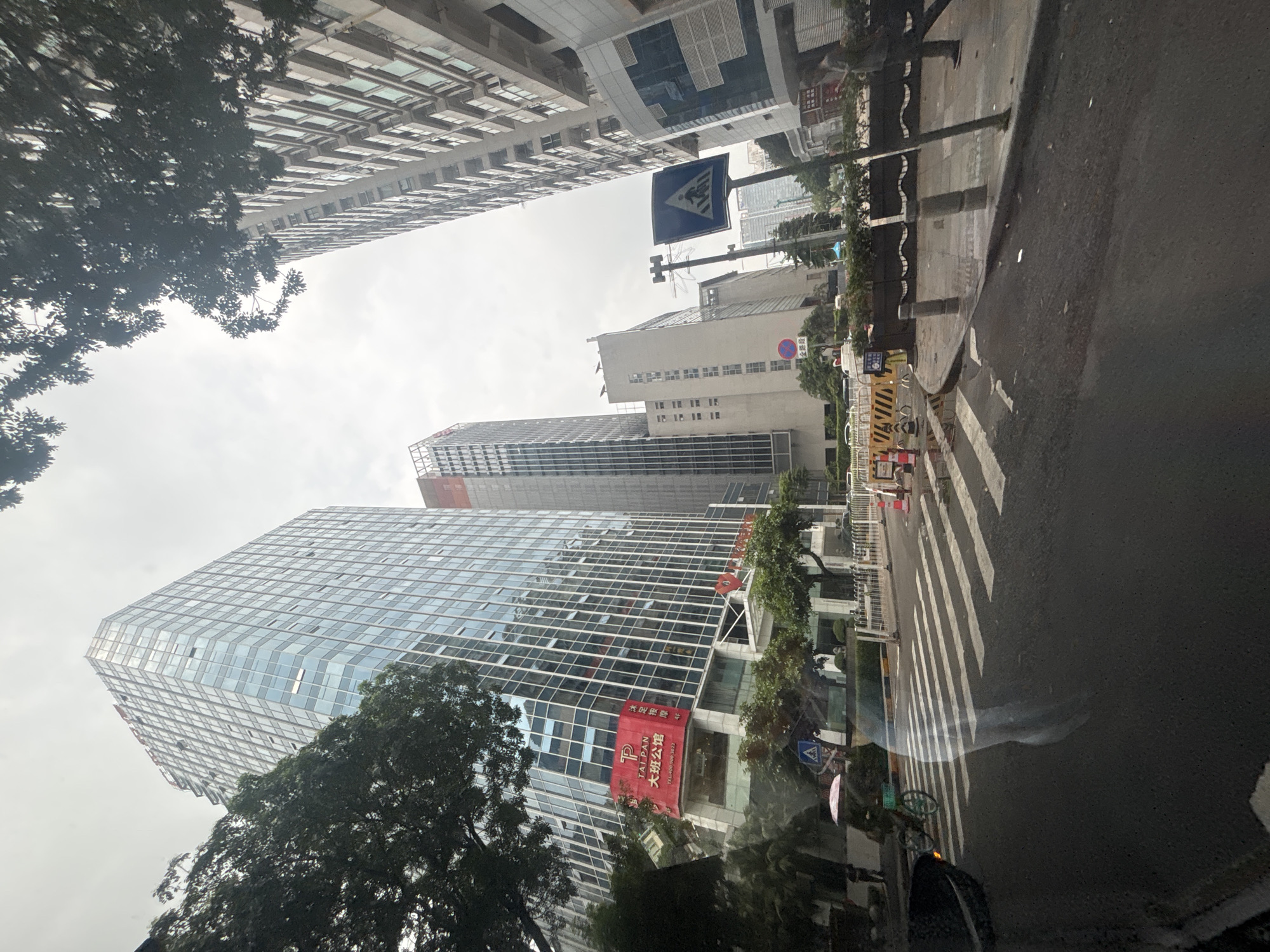 The image captures a street scene in Tianhe District, China, characterized by towering modern architecture and a busy urban environment. In the foreground, a wet asphalt road with a pedestrian crosswalk is visible, suggesting recent rain. A blurry, translucent reflection appears on the windshield of a vehicle, distorting the view of the road. To the left, a person on a bicycle with a pink umbrella navigates the street.
Dominating the background are several tall, glass-facaded buildings, typical of a commercial district. One prominent building features a large red sign with Chinese characters and the letters "TALPAN". To the right, another skyscraper displays a more traditional, lighter-colored facade. Traffic signs, including a pedestrian crossing sign, are present along the sidewalk, where a few individuals are walking. The sky is overcast, contributing to a muted light throughout the scene. Trees with dense foliage line parts of the street, adding a touch of green to the urban landscape. The overall impression is one of a bustling, contemporary city.