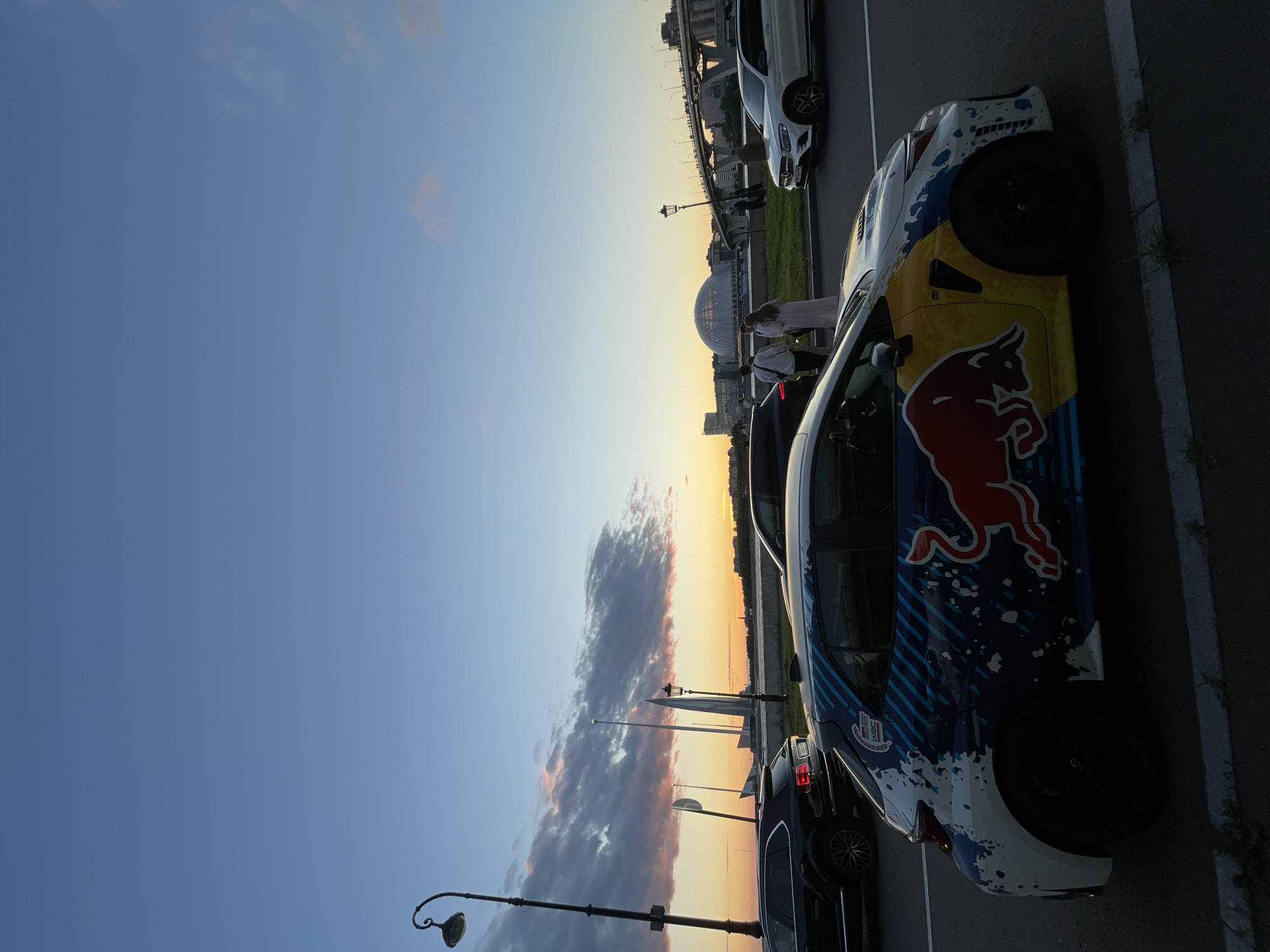 In an outdoor urban setting at dusk, a white hatchback adorned with a vibrant Red Bull livery and blue, white, and yellow paint splatters is parked on an asphalt road. The car's design includes the iconic Red Bull bull graphic and "GR YARIS WRC" branding. Several other vehicles are visible in the background, including a black SUV and a white sedan. A group of people, two women and a man, are walking on a grassy area behind a low wall. The skyline features a distinctive domed building and a modern skyscraper, possibly the Lakhta Center. A bridge with traffic and pedestrians spans a body of water in the distance. The sky exhibits a gradient of blue and pink, indicating sunset. A vintage-style lamppost stands to the left of the frame.