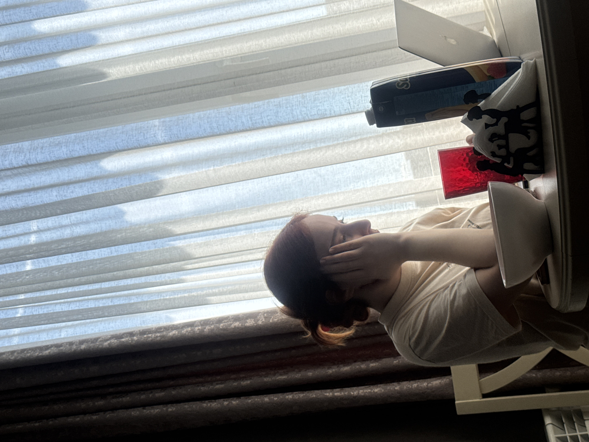 A person sits at a table in a room with a window covered by blinds. The person has their head in their hands, resting on their arms on the table. On the table is a laptop, a juice box, a decorative napkin holder with napkins, a red glass, and a plate. The blinds are partially open, revealing a view of the sky. The lower portion of the window is covered with a curtain. The location is likely Tbilisi, Georgia, although there are no specific visual cues to confirm this.