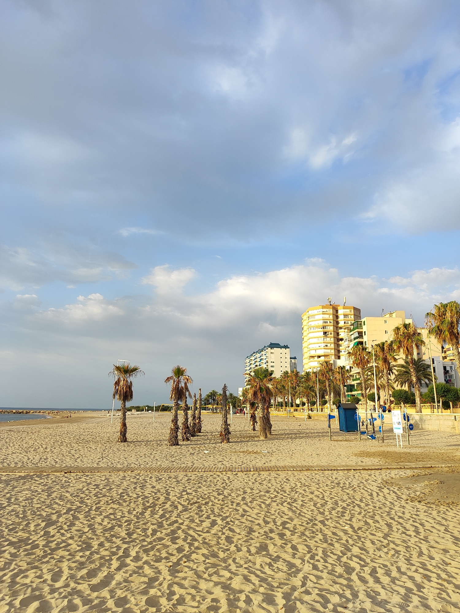 The image depicts a beach scene in El Campello, Spain, likely near the marina given the presence of buildings and palm trees. In the foreground, the sandy beach is textured with footprints and subtle shadows. A wooden walkway runs across the sand. Scattered palm trees, some with dry fronds, are visible, with a few taller ones to the right. In the middle ground, there are beach amenities, including a blue changing cabin and exercise equipment. Beyond that, the beach extends towards the sea, with a breakwater or jetty visible on the left. Several buildings, including apartment complexes and a distinctive yellow building, line the background, interspersed with more palm trees. The sky is partly cloudy with a mix of white and gray clouds. There are a few distant figures on the beach and near the water's edge.