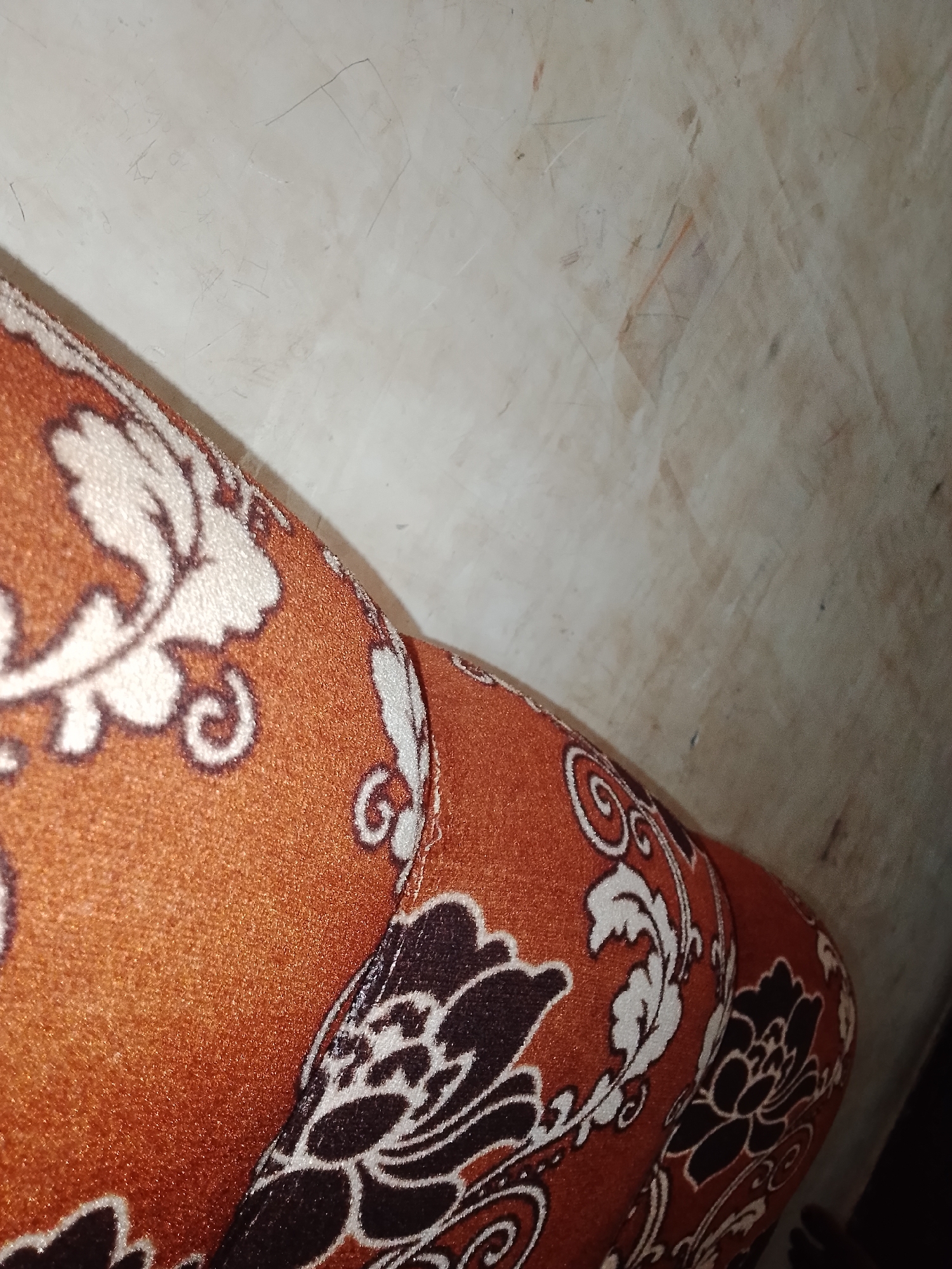 The image is a close-up shot of a patterned fabric, likely upholstery or clothing, with an orange base color and a floral design in white and dark brown. The fabric has a textured appearance. In the background, a wall is visible, appearing to be made of a light-colored, possibly concrete or plastered material, with some faint markings or scuffs. The lighting suggests an indoor environment. The scene is devoid of any people or specific landmarks that would indicate a precise location within Jalingo, Nigeria.