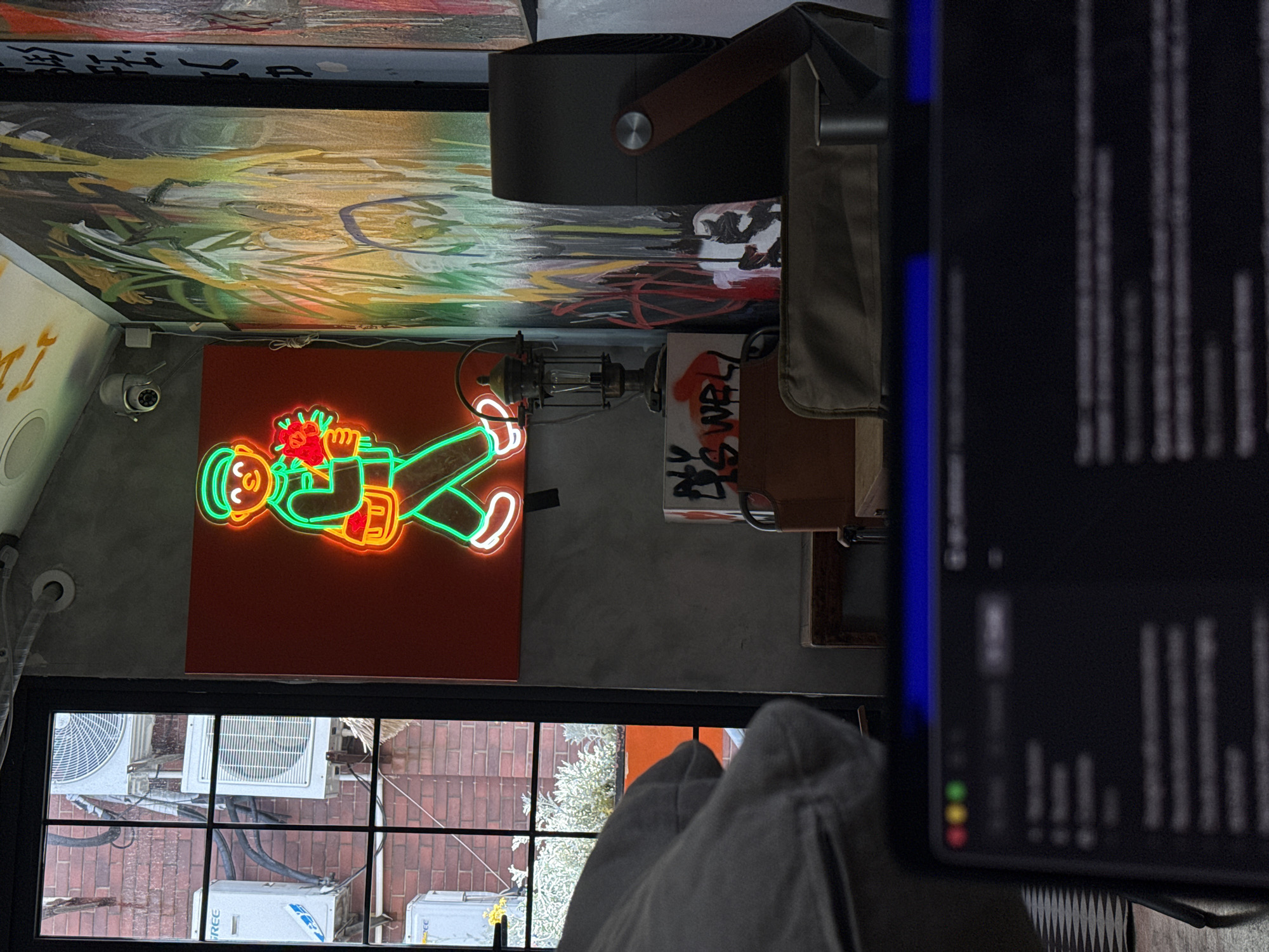 The image captures an indoor scene, likely in an urban setting within Putuo District, China, viewed from a slightly elevated perspective. The overall impression is that of a trendy, artistic space, possibly a cafe or studio, illuminated by artificial light, suggesting evening or nighttime.
The focal point is a vibrant neon sign on a red wall, depicting a stylized figure with a green outline, orange face and hands, and holding a red object that resembles a heart or flowers. To the left of the sign, a security camera is mounted on the wall. Above, the ceiling is adorned with an abstract, colorful mural featuring brushstrokes in yellow, green, black, and red hues.
To the right of the neon sign, a dark, modern-looking air purifier or fan with a brown leather-like handle is visible. Below it, an industrial-style lantern with an exposed filament bulb adds to the decor. A wooden chair or table is partly visible, bearing graffiti that reads "YS WELL".
Below the neon sign, a window looks out onto an exterior scene with multiple outdoor air conditioning units, including one branded "GREE," and a red brick ground below. Part of a person's leg, clad in grey textured fabric, is visible in the foreground, partially obscuring the view through the window.
On the far right, a blurred electronic screen, possibly a tablet or monitor, dominates the foreground. It displays vertical lines of unreadable text or code on a dark background, with a prominent blue edge and small red, yellow, and green circular indicators at the bottom left.