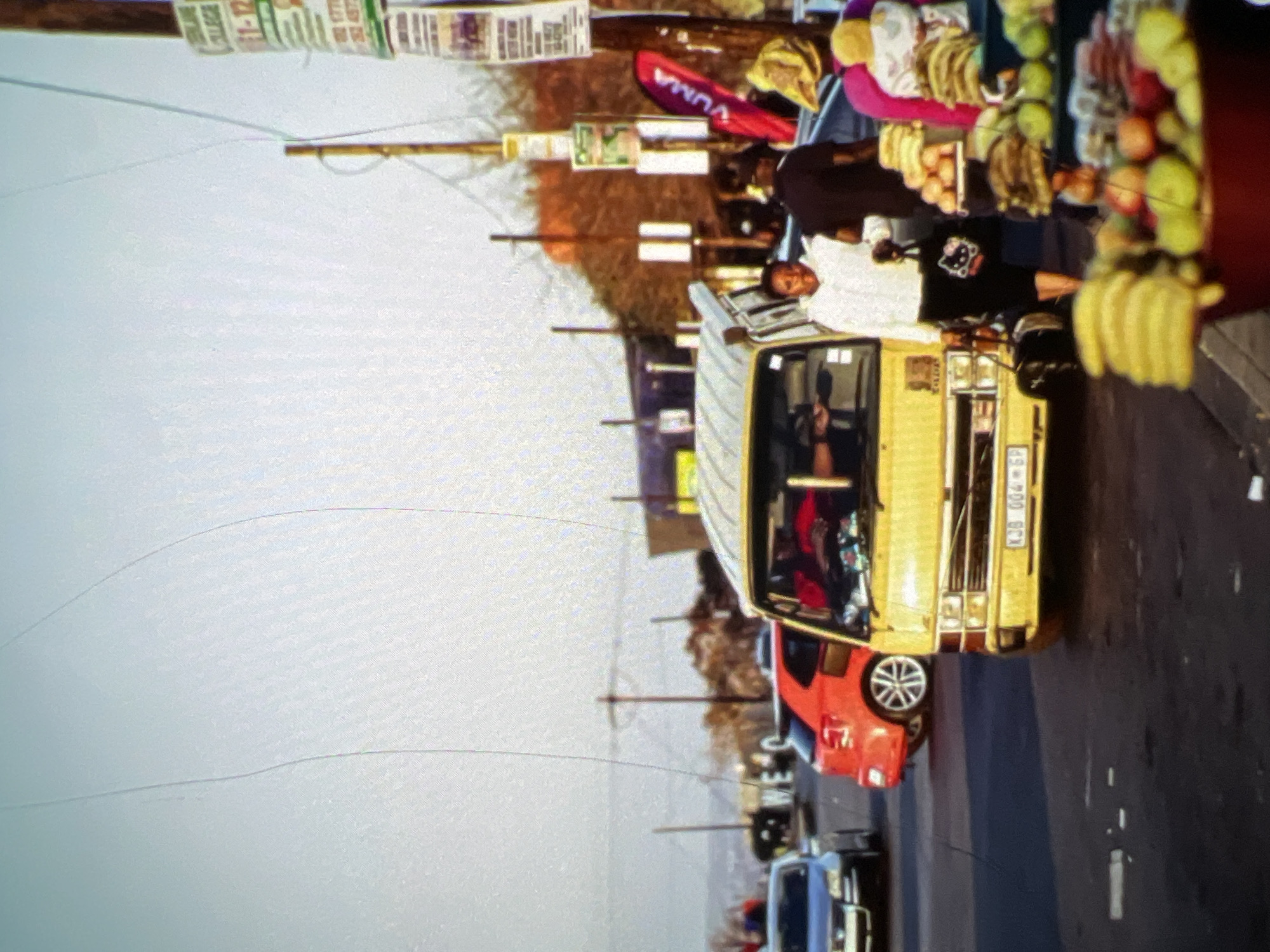A street scene in Lagos, Nigeria, features a yellow minibus taxi prominently in the foreground, its front license plate reading "XJ8 004 GP". A woman in a white top and black skirt, carrying a black bag with a Hello Kitty design, walks beside the taxi. Behind her, a man in a dark shirt and a woman in a pink and yellow headwrap are visible, along with a fruit stall displaying bananas, apples, and other produce. Other vehicles, including a red car and a silver car, are present on the road. Overhead wires and utility poles with posters are visible, suggesting an urban environment. The sky is overcast.