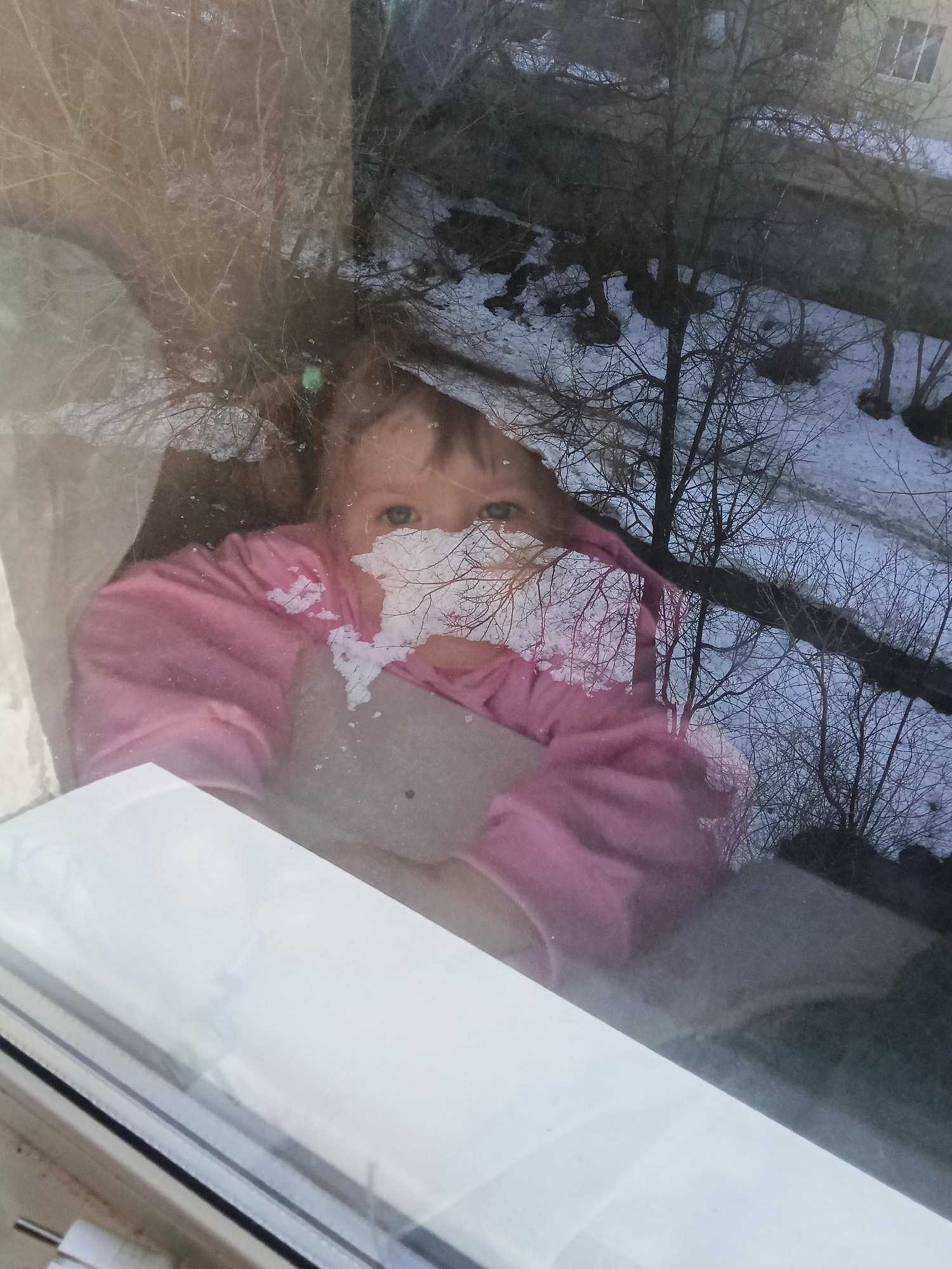A young child in a pink garment is visible through a window, seemingly looking outside. The reflection in the window shows trees with bare branches and patches of snow on the ground, indicating winter conditions. A building with a window is also reflected. The location is plausibly Chaikovsky, Russia, as per the request. The child is holding a flat, light-colored object. The window frame is white.