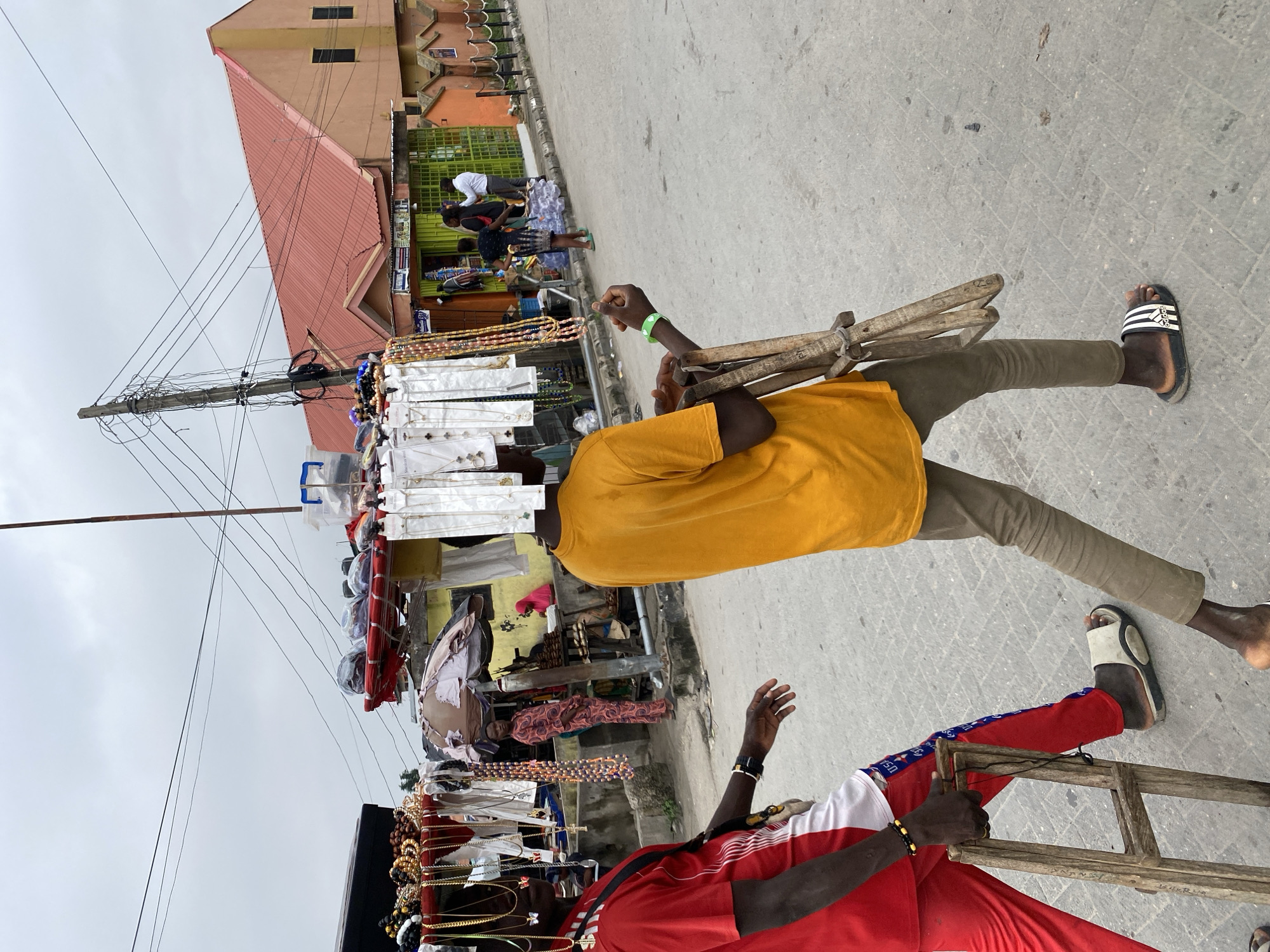 Two men walk along a paved street in Ado, Nigeria, each carrying a display of merchandise balanced on their heads. The man in the foreground wears a red and white shirt and red pants. The other man wears a yellow shirt and khaki pants. Both wear sandals. The displays include jewelry, necklaces, and other accessories. A building with a red roof and a utility pole with numerous wires are visible in the background, along with other people browsing merchandise displayed along the side of the street.