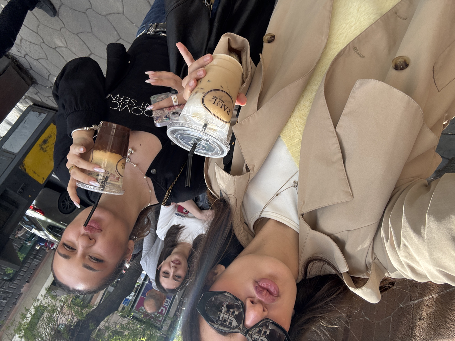 The image, taken in Yerevan, Armenia, captures three women enjoying iced beverages outdoors on a bright, sunny day, likely during a casual gathering or outing. The photograph is taken from a slightly upward and rotated angle, presenting a candid moment.
In the immediate foreground, a woman on the right is seen from the chest up, sporting dark sunglasses that reflect a building, and light pink-toned lips. She is dressed in a beige trench coat layered over a white top and a yellow knitted sweater. Around her neck, a delicate silver necklace with a small charm is visible. She holds a clear plastic cup filled with an iced coffee, clearly displaying the brand "PAUL" and the text "depuis 1889".
Beside her, to the left in the mid-ground, another woman is partially visible. She is wearing a black top adorned with some text across the chest that appears to read "LOVIC SERVIC" or "LOVIC SERVT" with decorative elements. Her hands, adorned with rings and long, light-colored manicured nails, hold a second clear plastic cup with an iced drink. A beaded necklace is visible around her neck. She looks towards the camera with a playful, slightly pursed lip expression.
In the background, a third woman with dark hair is partially seen between the two, wearing a white top and smiling subtly.
The setting suggests an urban outdoor environment, with elements like pavement, glimpses of parked cars, and some green foliage visible behind the subjects. The bright sunlight indicates it is daytime, and the casual attire, including trench coats and iced drinks, implies pleasant weather. The overall impression is one of relaxed camaraderie among friends.
