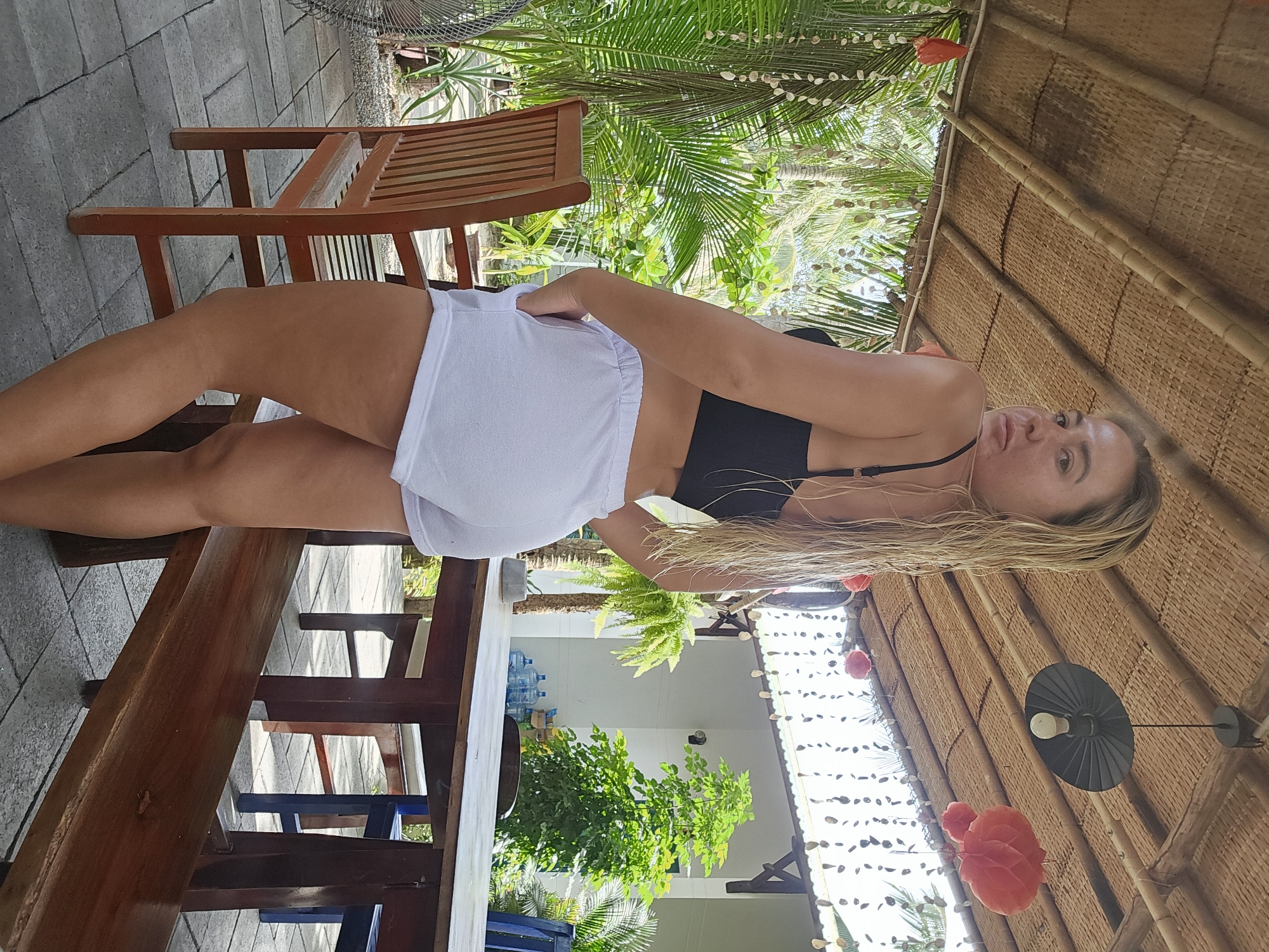 A young Caucasian woman stands outdoors on a tiled patio, looking over her shoulder at the camera. She has blonde, wet hair and is wearing a black bikini top and white shorts. Her hands are in her pockets. Behind her, a wooden table and chairs are set up under a thatched roof. Lush green foliage, including palm trees, surrounds the patio. The lighting suggests it is daytime. The woman's pose and attire suggest a casual, tropical setting. The scene does not provide any information to suggest it is located in Phu Thuy Ward, Vietnam.