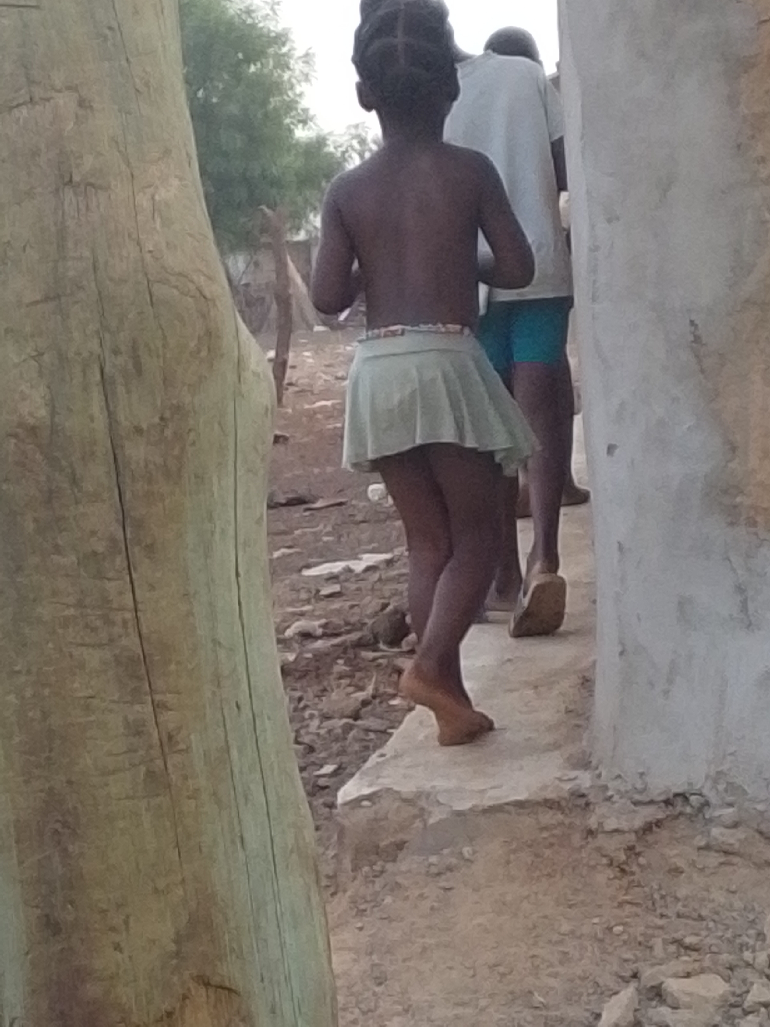 The image shows two figures, viewed from the rear, walking on a raised concrete path in an outdoor setting. A young girl with braided hair is in the foreground, wearing a light green skirt and no shirt. An older person, wearing a white shirt and blue shorts and sandal-type shoes, walks behind her. The ground is dirt with scattered rocks and debris. To the left of the figures, a large tree trunk is partially visible. A concrete wall stands to their right. Trees are visible in the background. The city of Tamale, Ghana is not discernible from the image.