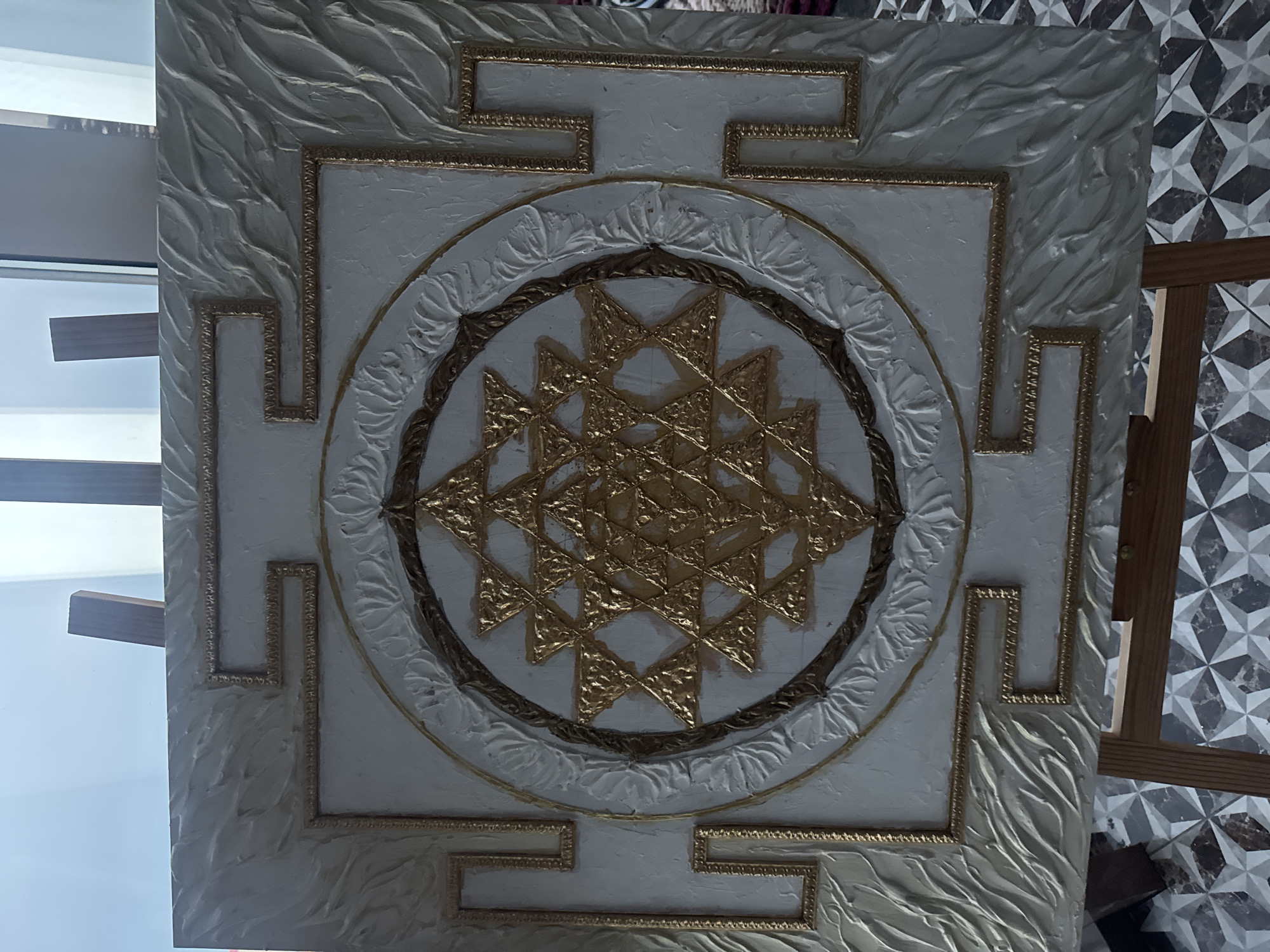The image depicts a square, framed artwork featuring a complex geometric design. The background is white with textured, swirling patterns. A square frame surrounds the central design, accented with a Greek key pattern in gold. Inside the square is a circular design, also with gold trim and a leaf-like motif. At the center of the circle is a gold geometric pattern composed of intersecting triangles. The background of the central design is white.
There is no indication of a location in the city of Novosibirsk, Russia, in this image. The surrounding environment includes a window with a view and a wooden easel stand. The floor tiles have a geometric pattern.