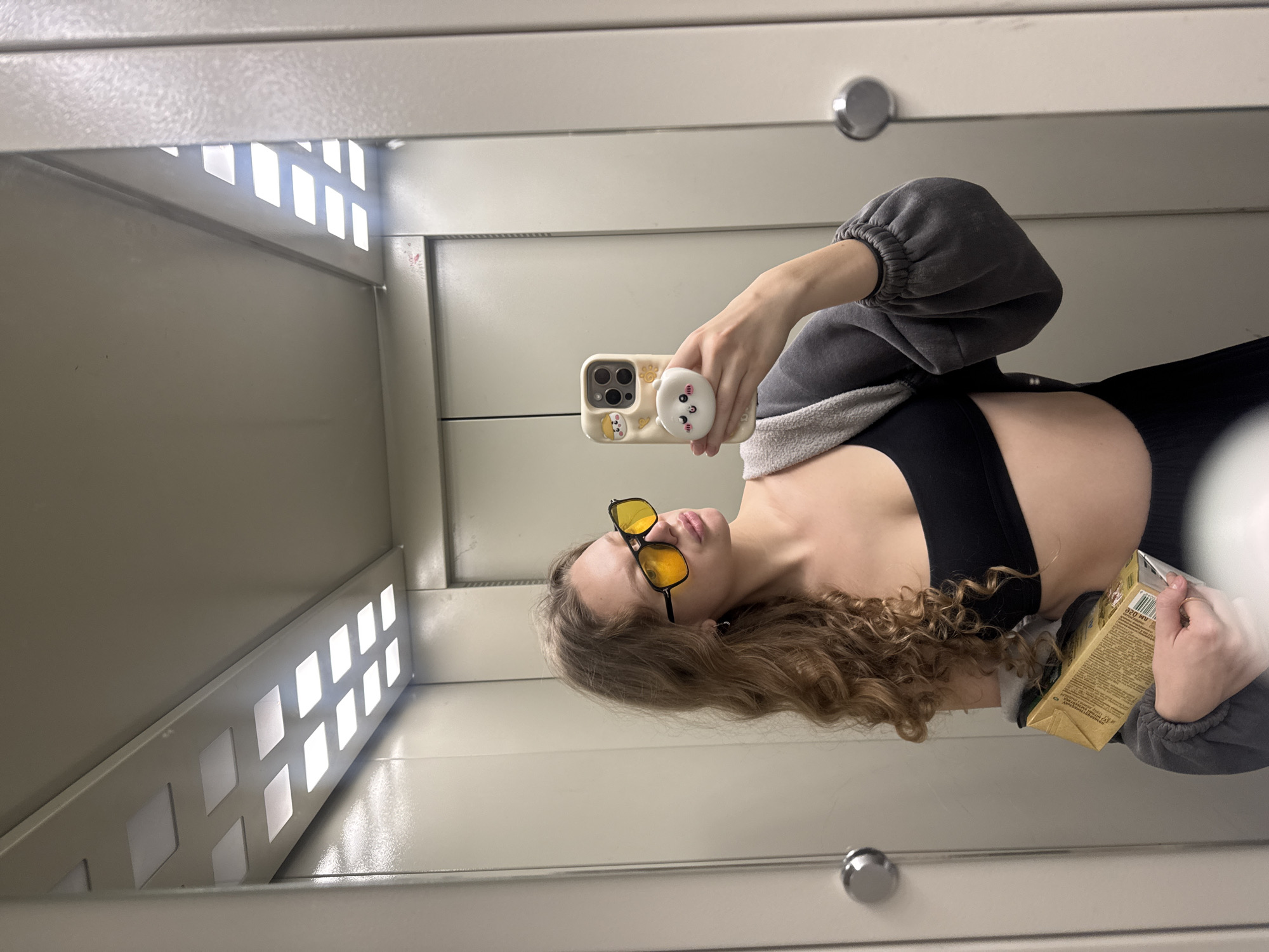 A young woman with long, curly blonde hair is taking a mirror selfie in what appears to be an elevator. She is wearing yellow-tinted sunglasses, a black sports bra, and a grey hooded sweatshirt worn open. She is holding a small, rectangular golden box in her left arm and her phone in her right hand, which has a case with cartoon characters and a white pop socket. The elevator walls are a neutral beige color, and the ceiling features a row of rectangular fluorescent lights. The overall environment suggests an indoor urban setting, possibly in Izhevsk, Russia, based on the provided context.