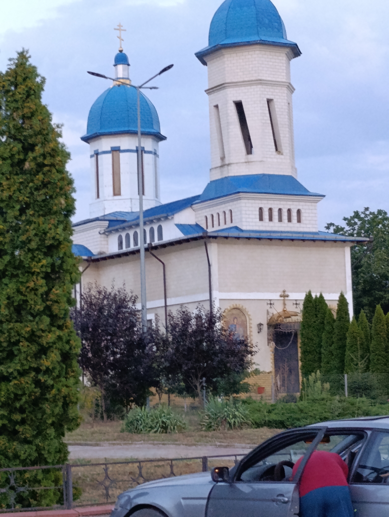 In the city of Cocieri, Moldova, a multi-story building with a blue domed roof and white facade stands prominently. The building features multiple windows and architectural details indicative of a church. In the foreground, a silver car is parked, and a person wearing a red shirt and blue pants is partially visible inside the driver's side, with the door ajar. Trees and bushes surround the building, and a fence is present in the lower portion of the image. The sky is overcast.