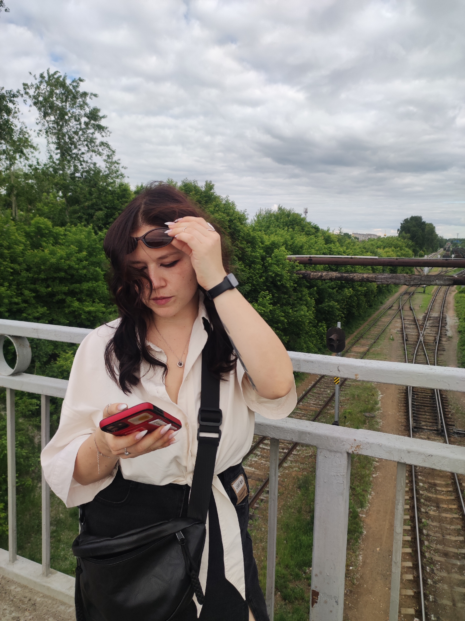 A young woman with dark hair is standing on a bridge over railroad tracks in Chaikovsky, Russia. She is wearing a white open-collar shirt, black pants, and a black crossbody bag. She is holding sunglasses in her right hand and looking at her red smartphone with her left hand. Her smartwatch is visible on her left wrist. The background shows lush green trees and a cloudy sky. The railroad tracks split into multiple paths, suggesting a junction or switchyard.