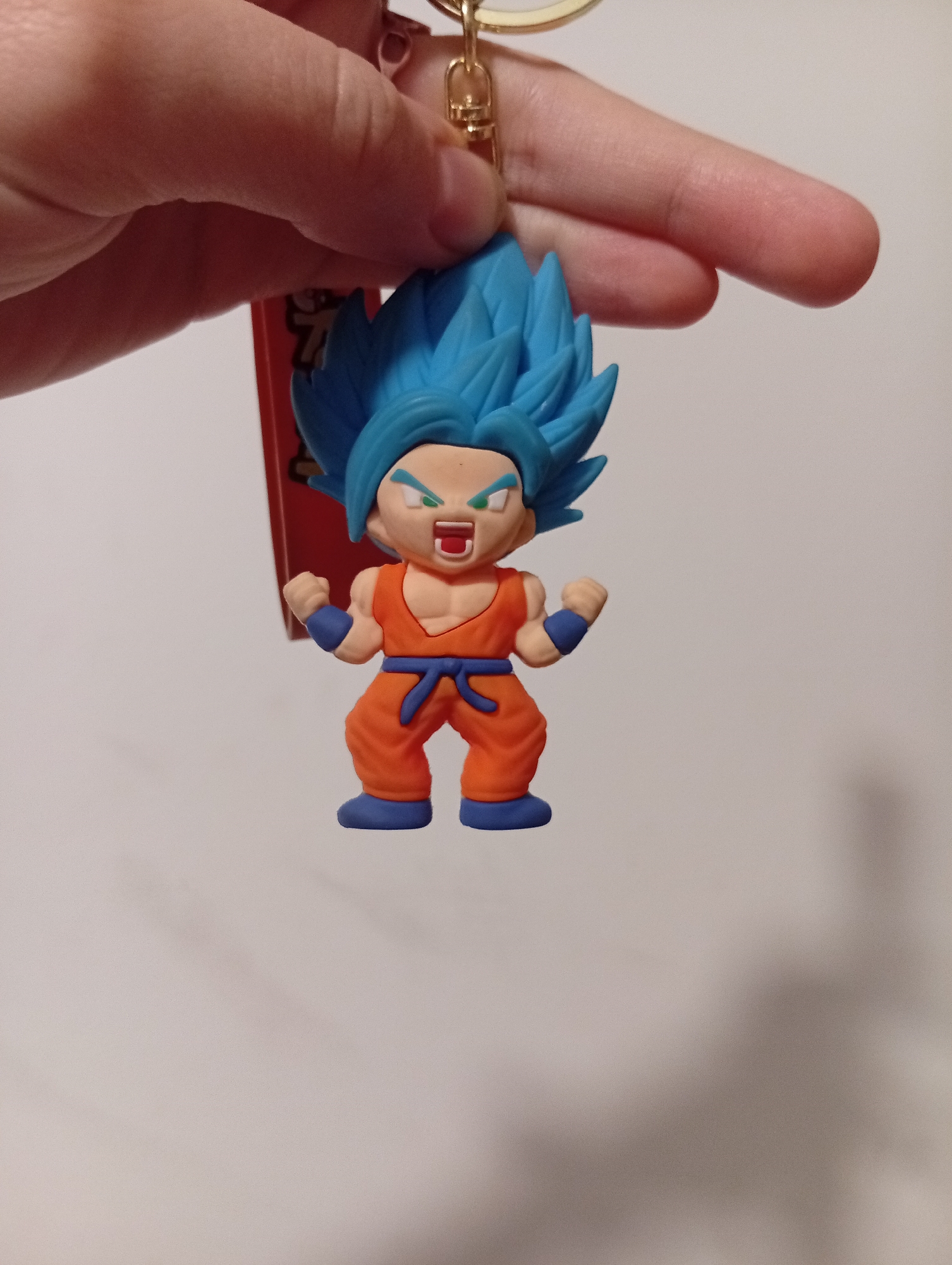 A close-up shot captures a chibi-style keychain figurine of Goku from Dragon Ball, depicted in his Super Saiyan Blue form, held aloft by a human hand. The figurine is crafted with blue, spiky hair and features an orange gi, blue belt, blue wristbands, and blue boots. Its face shows an aggressive expression with an open mouth revealing teeth and green-colored eyebrows and eyes. Both of its fists are clenched, adding to the powerful stance.
The figurine is attached to a golden metal keyring clasp, which is being held by a fair-skinned hand. Behind the figurine, a red tag is partially visible, likely part of the product's packaging or branding, though any text on it is too blurry to decipher. The background is a plain, light-colored surface, possibly a wall, appearing out of focus. The overall impression is a detailed, collectible item. The image's context is Buenos Aires, Argentina.