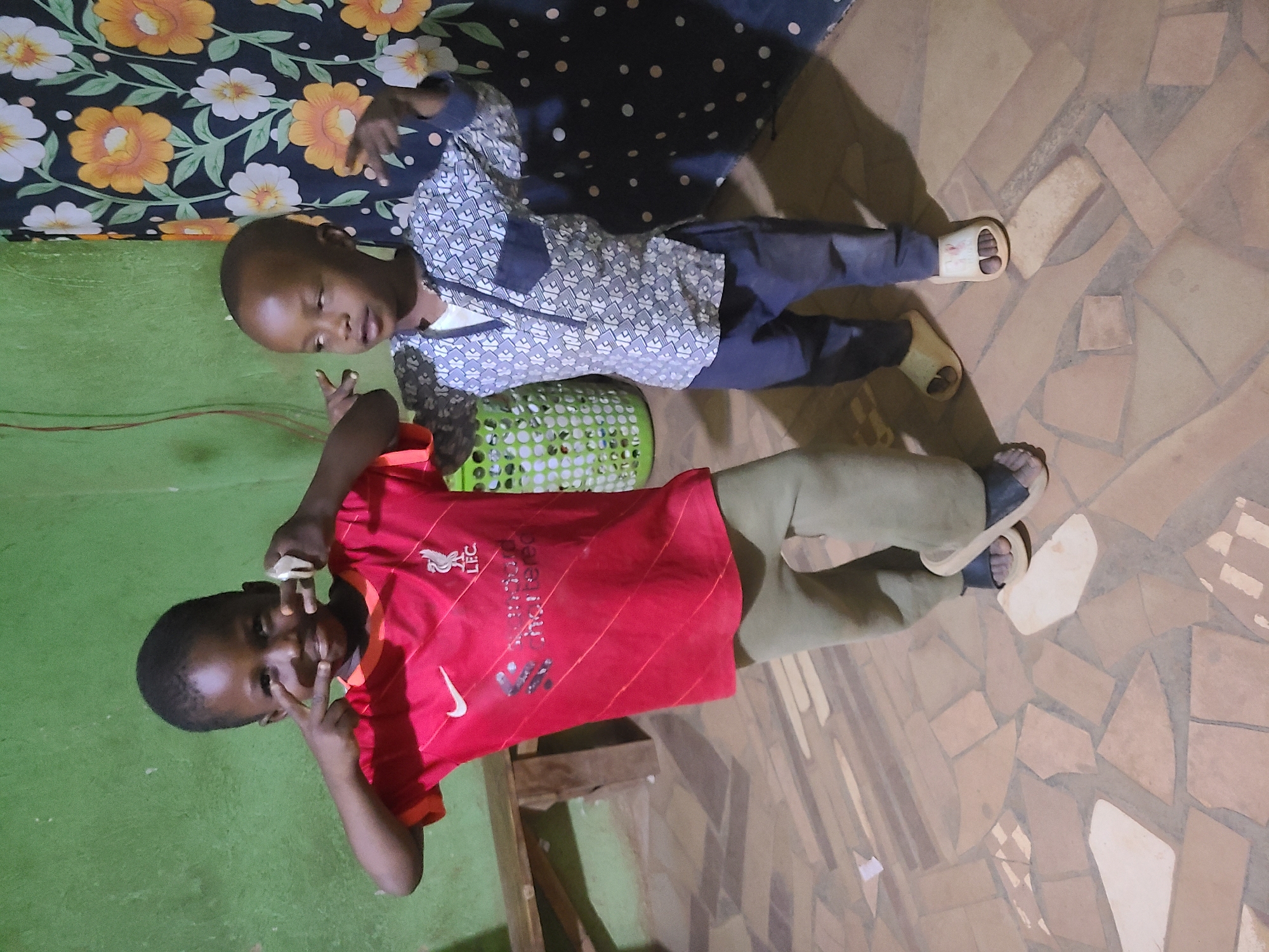 Two children are lying on the floor of a room in Unknown, Unknown. The child at the top is wearing a patterned shirt and dark pants. The child at the bottom is wearing a red sports jersey and khaki pants. Both are making a peace sign with their hands. A green basket sits between them. The floor is tiled with beige and white tiles. A green wall is visible on the left side of the frame and a floral patterned blanket is visible at the top.