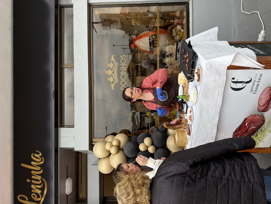 An outdoor scene captured in front of the "Leninha" confectionery in Felgueiras, Portugal, depicts a food tasting event.
A female vendor, with reddish-brown hair and glasses, wearing a salmon-colored shirt, brown apron, and blue gloves, stands behind a white-draped table. She gestures with her right hand while addressing two female customers. The customer in the foreground, seen from the back, wears a black quilted jacket. The second customer, slightly behind her, with brown hair, is tasting a food sample from a toothpick.
The table features a variety of food items: a leg of cured meat on a wooden stand, multiple plates with sliced meats and cheese samples served with toothpicks, a block of cheese, and a small green bottle. On the right side of the table, an electric griddle contains cooking slices of meat. The front of the table is adorned with a banner promoting "Quinta dos Fumeiros," showcasing images of smoked meats. A white power strip with a cable is visible on the ground to the right, powering the griddle.
To the left of the table, a beige and black balloon cluster stands. Behind the vendor and the table is the storefront window of "DOCINHOS Helena Gonçalves - Docaria Tradicional," displaying an oversized Easter egg decoration and ceramic bunny mugs, indicating an Easter theme. The store's primary sign, "Leninha," in gold script, is mounted on the black fascia above the window. The sky, reflected in the window, appears overcast.