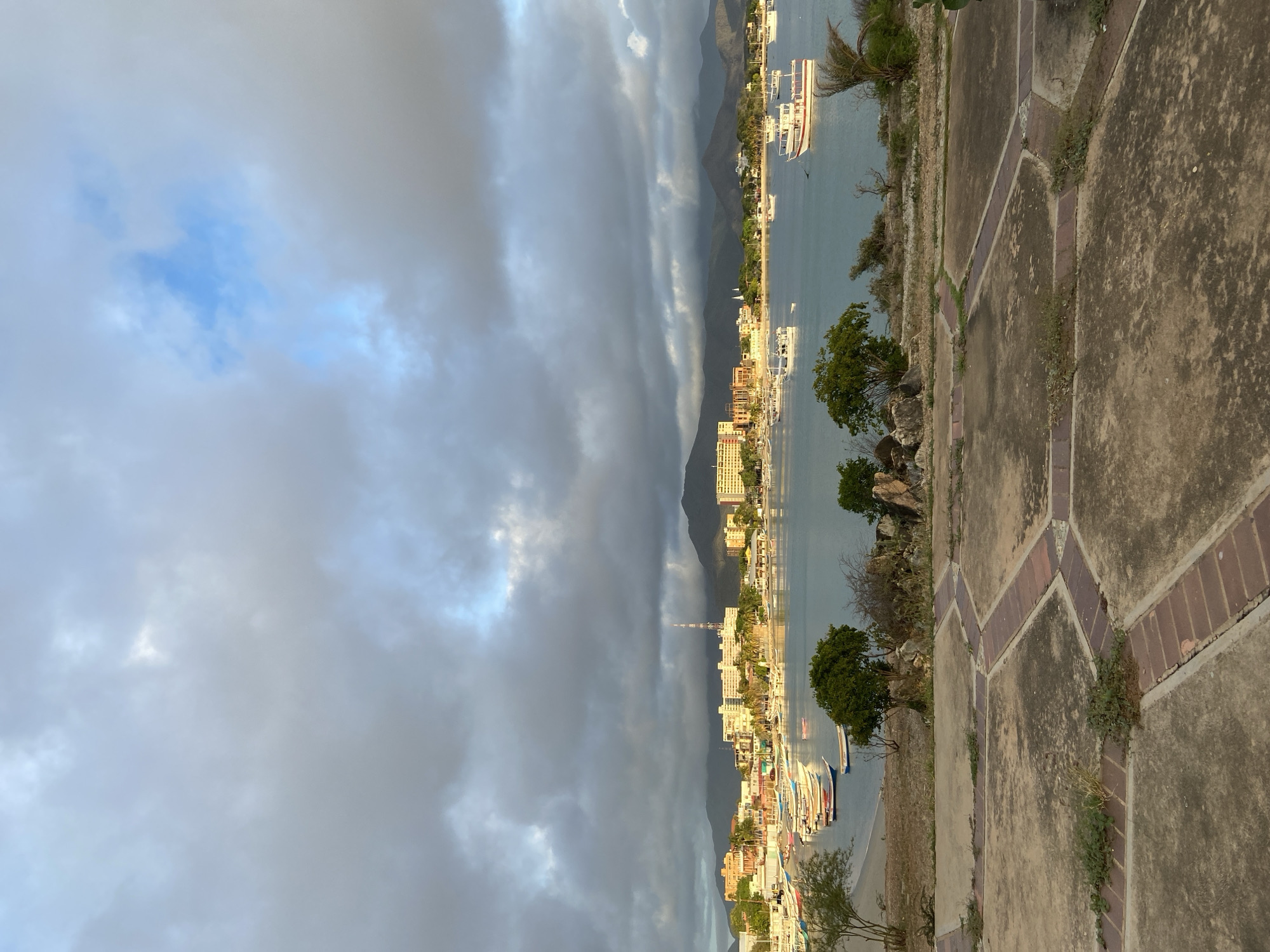 This is a photograph of the bay in Juan Griego, Venezuela, taken from an elevated viewpoint. The foreground features a paved area with a decorative brick pattern, overgrown with sparse vegetation. Beyond this, the scene opens to a tranquil bay filled with numerous boats of various sizes, predominantly small fishing vessels and larger tour boats, anchored or gently moving on the water.
The middle ground showcases the coastline of Juan Griego, characterized by a sandy beach lined with a collection of buildings, including hotels, apartment complexes, and smaller structures. Palm trees and other lush vegetation are visible among the buildings. A prominent radio tower stands tall on the horizon, to the left of center.
In the background, a range of mountains forms a natural backdrop to the city and bay. The sky is dramatic, filled with a mix of heavy, dark clouds and patches of bright blue, suggesting an overcast but potentially clearing day. The lighting indicates it may be late afternoon or early evening, with sunlight casting a warm glow on parts of the scene. No people are explicitly visible in the image.