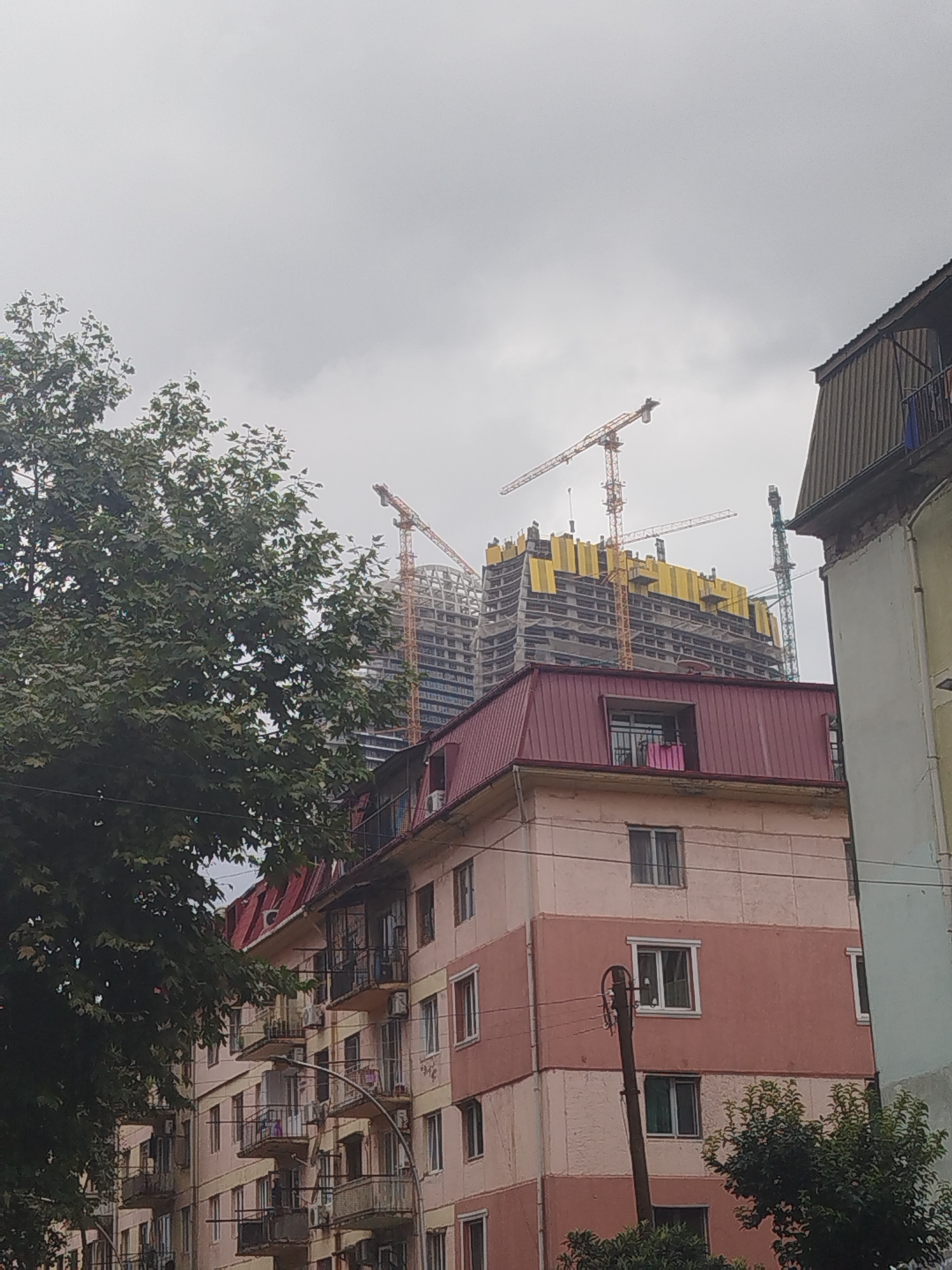 This image captures a view of urban development in Batumi, Georgia, characterized by a mix of existing residential buildings and a large construction site. Dominating the background is a massive, partially constructed building, its concrete frame exposed and clad in yellow sheeting. Several prominent yellow construction cranes are actively engaged in the building process, reaching high into the overcast sky. To the left, a large, leafy green tree partially obscures the view, its branches extending across the frame. In the foreground, several older, multi-story residential buildings are visible. These buildings feature balconies with railings, windows, and visible air conditioning units, suggesting a lived-in urban environment. The overall atmosphere is one of ongoing development, with the new construction contrasting sharply with the established residential structures.