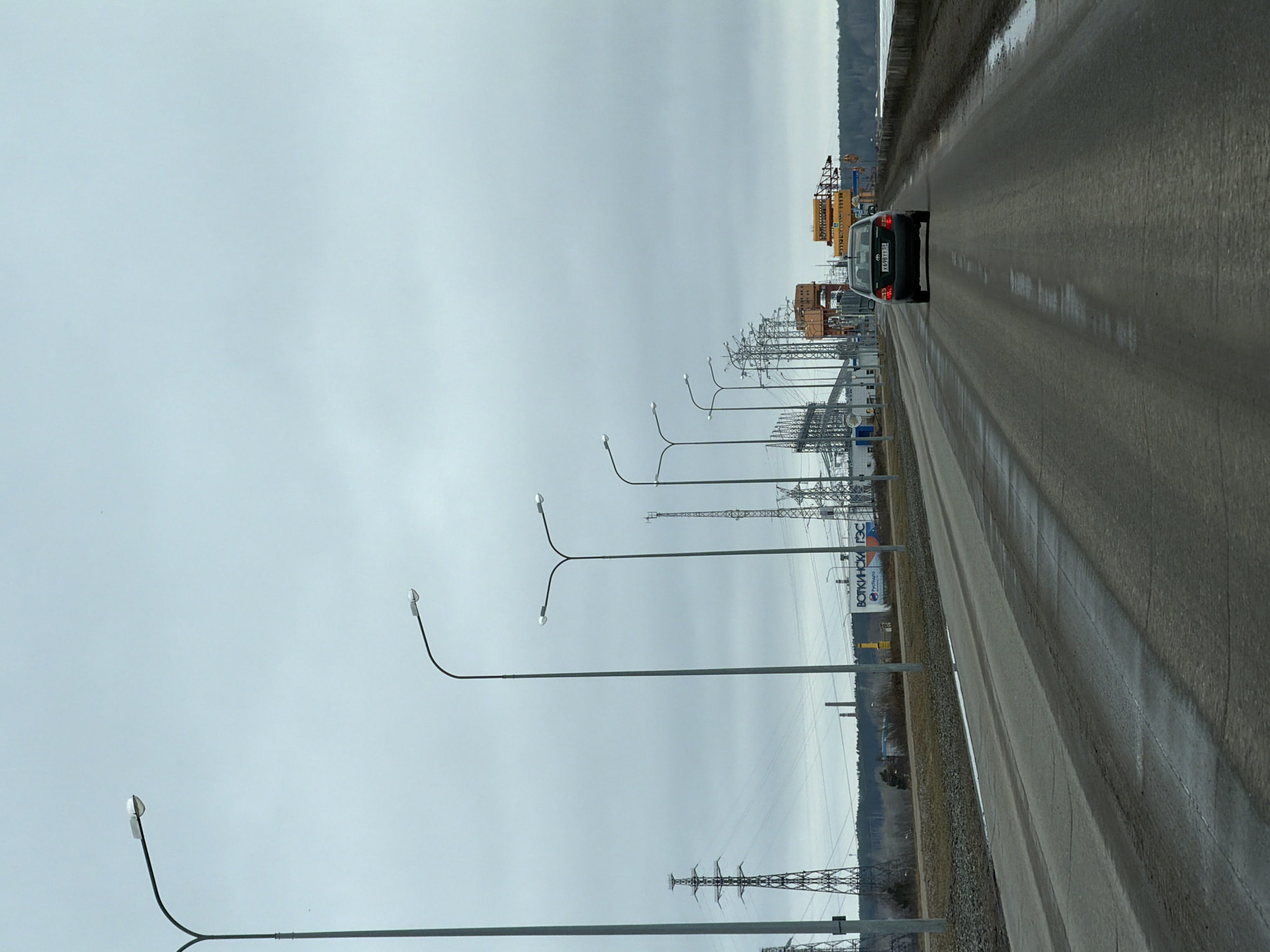The image depicts a road scene in what is likely Chaikovsky, Russia, given the Cyrillic text visible on a sign along the roadside. The road is wet, suggesting recent rainfall. A dark-colored car is traveling on the road, away from the camera. The landscape to the right of the road features a river or large body of water, with trees visible in the distance. Power lines and electrical infrastructure are prominent on the left side of the road, interspersed with streetlights. The sky is overcast.