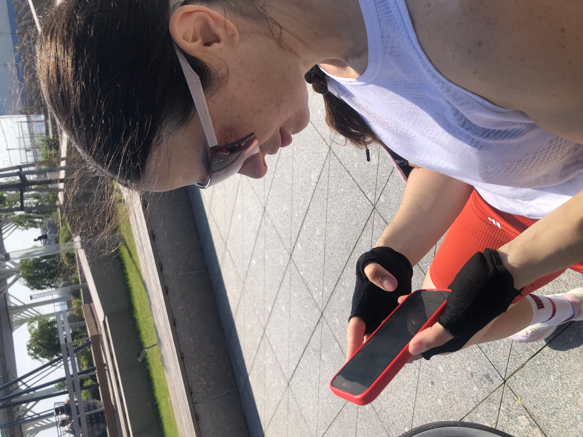 A medium shot captures a young woman with dark hair, wearing sunglasses and fingerless gloves, looking down at her red-cased smartphone. She is dressed in a white sleeveless top and red athletic shorts. The background suggests an outdoor urban setting with a paved ground, a grassy area, and some distant structures. The lighting indicates it is a bright, sunny day. The woman is seated, with a portion of a bicycle visible in the lower left corner of the frame. The environment appears to be a public space, possibly a park or waterfront promenade. The specific location in Saint Petersburg, Russia, is not identifiable from the visual cues present in the image.