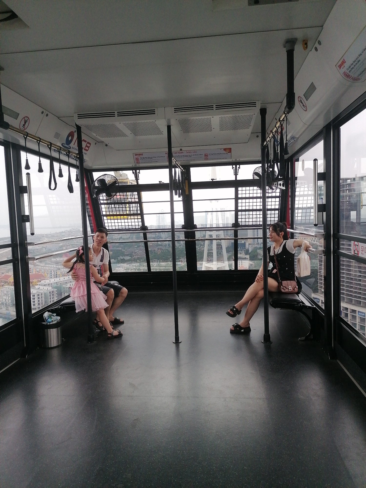 The image shows the spacious interior of a modern cable car cabin, providing a panoramic view of a cityscape and a body of water, indicative of a coastal area like Bai Chay Ward, Vietnam. The sky outside appears overcast, suggesting a cloudy day.
Three individuals are seated within the cabin, seemingly enjoying the scenic ride. On the left, a man in a white polo shirt and dark shorts is seated on a bench, his arm around a young girl who is sitting on his lap. The girl wears a pink dress and a matching hair bow. They are both wearing sandals. Opposite them, on a separate bench, a woman sits in a relaxed posture. She is dressed in a white t-shirt layered under a black top or dress, dark shorts, and dark platform sandals. She holds a plastic bag and is looking towards the man and girl with a gentle smile.
The cabin features large windows that offer expansive views of numerous urban buildings, some towering, and a distant body of water. The interior design is functional and clean, with a dark, speckled floor, white ceiling panels, and black metal framework. Overhead, there are handrails with hanging grips, and two circular fans are mounted on the side walls. Ceiling vents are also visible, likely for ventilation or air conditioning. A small metal trash can is placed on the floor near the man and girl.
Visible text includes "NATIONAL" and "ACB" logos, and a "SUNWORLD" logo, likely identifying the cable car operator. Safety instructions are posted in Vietnamese, with the heading "MY AN TOÀN | Safety rules", and phrases like "gừng dòng mo nủa cabin" and "Gia cười được đong mo tự động, vui lòng," which typically advise against leaning on automatic doors or bringing outside food/drinks. The overall impression is one of a calm, leisurely journey.