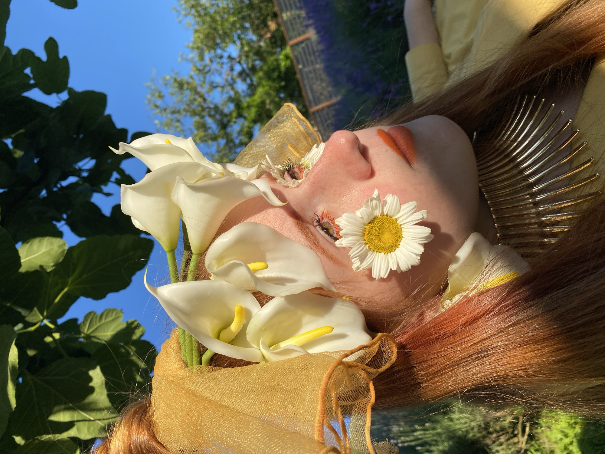 The media file is a close-up, horizontally oriented photograph of a person's face, adorned with flowers and metallic accessories, set against an outdoor backdrop. The subject, with reddish-brown hair, appears to be lying down or positioned such that their face is tilted upwards. Their eyes are closed, and their lips are painted a striking orange.
Several white calla lilies and a single white daisy are artfully arranged on the subject's face, particularly around the eyes and nose. A gold, mesh-like fabric is visible, draped around the neck and possibly serving as part of an elaborate headdress or garment. A metallic, fan-shaped accessory gleams behind the subject's head. The background reveals a bright blue sky with soft clouds, green foliage from trees and plants, and a blurred hint of a structure, possibly a fence or trellis, in the distance. The lighting suggests it is daytime, likely sunny, casting shadows and highlighting the textures of the flowers and the subject's skin.
The overall impression is one of ethereal beauty and artistic expression. The arrangement of flowers and the metallic elements create a fantasy-like or artistic portrait. The subject's closed eyes and gentle expression evoke a sense of peace or serene contemplation. The location context of Verkhneye Uchdere, Russia, combined with the lush greenery, suggests a natural, possibly garden or park setting. There is no visible text in the image.