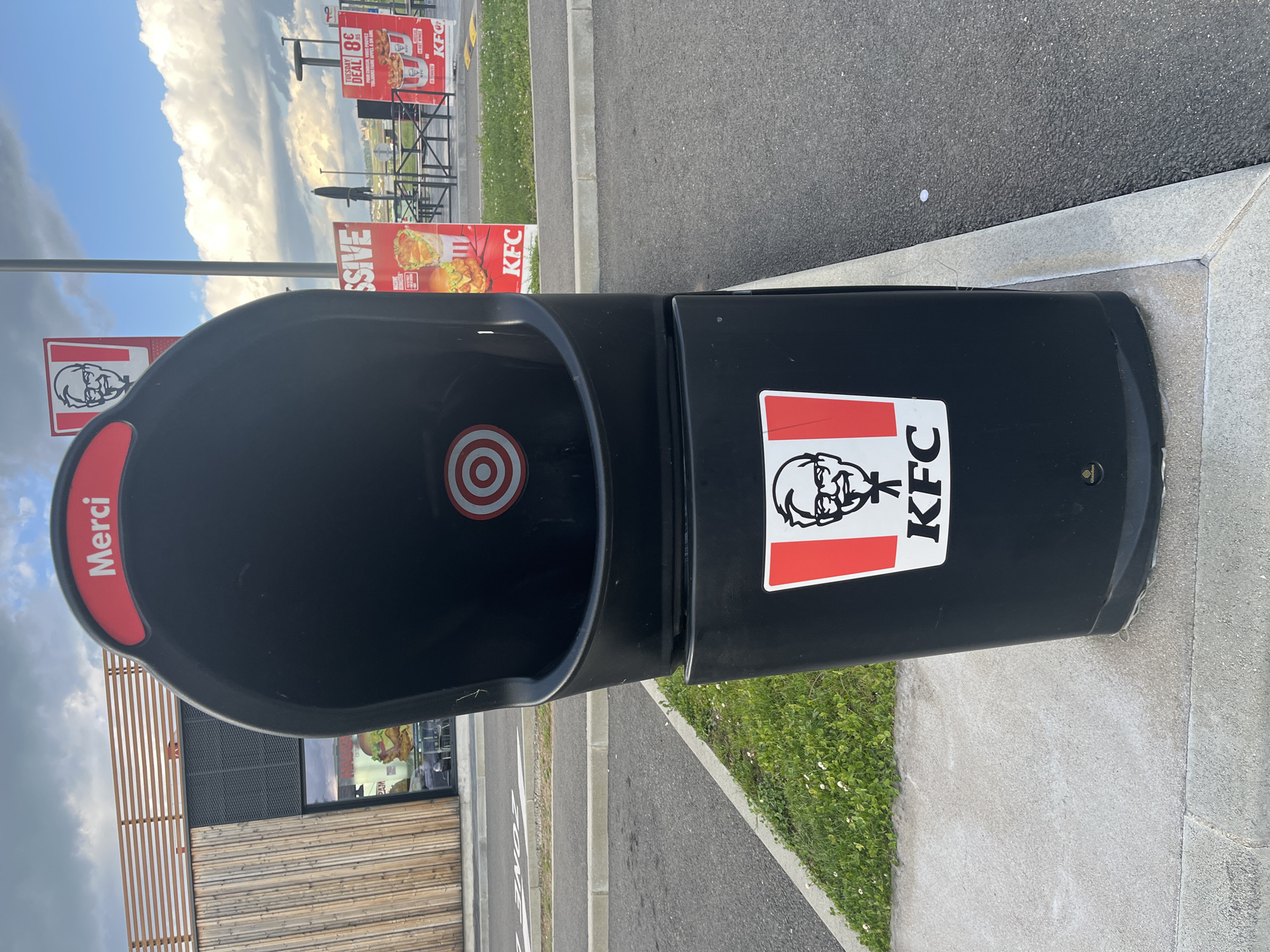 The image shows a black KFC trash receptacle in Andrézieux-Bouthéon, France. The bin has a red stripe with "Merci" printed on it, a red and white target on the interior, and the KFC logo on the side. In the background are KFC signs, the restaurant building, and the sky. The trash receptacle is located on a concrete pad, adjacent to a paved road. A grassy area borders the pad.