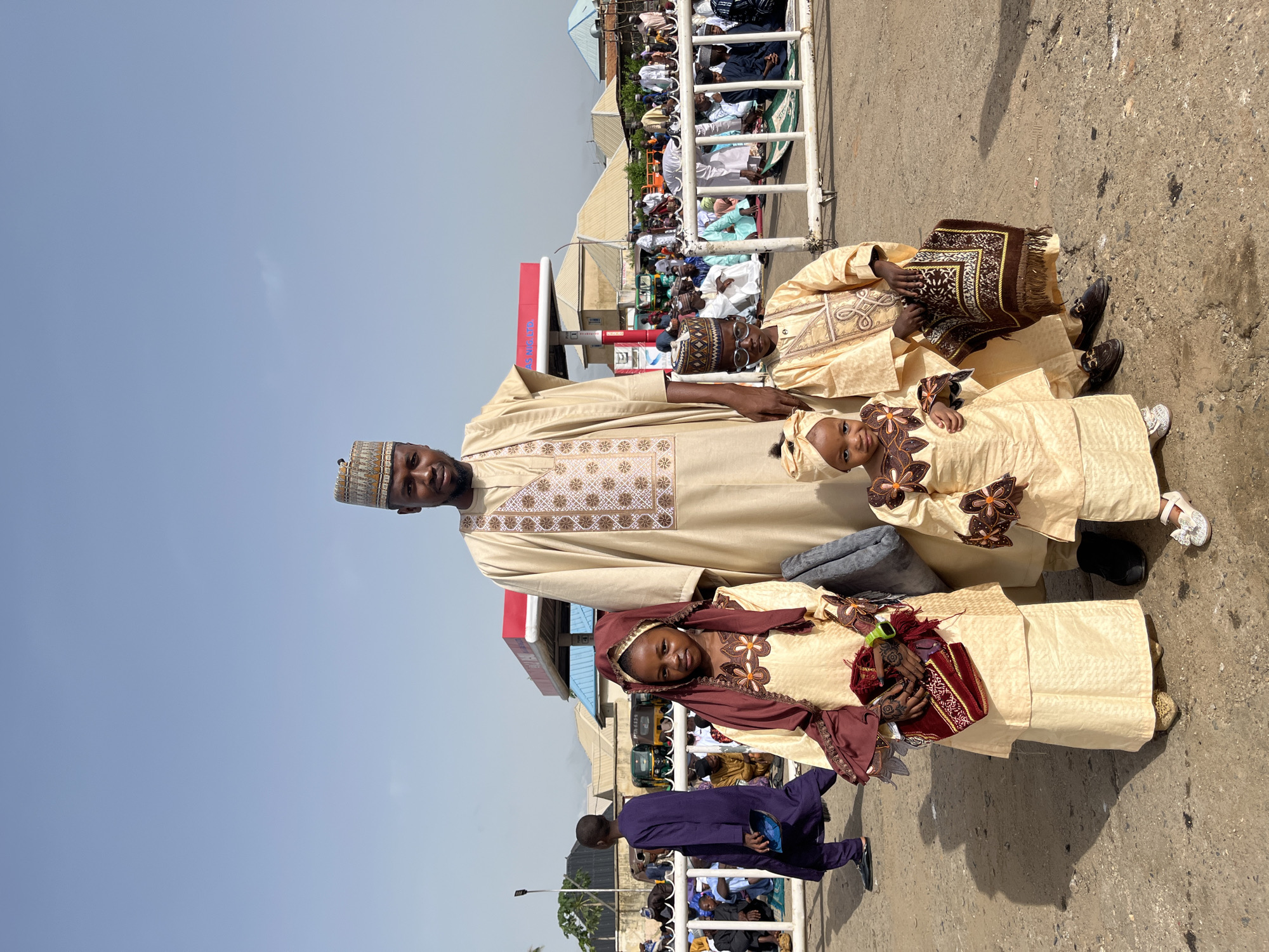 The image shows a man and two young girls standing outside, posing for a photo. The man is wearing a long, light-colored robe with decorative patterns and a matching cap. The girls are dressed in similar outfits with decorative accents. One girl holds a small bag. They are standing in front of a crowd of people seated on the ground behind a white metal barrier. In the background, there are buildings, signs, and a gas station. The photo appears to be taken outdoors on a sunny day. The location is identified as Jos, Nigeria, based on the presence of an "AS Nig LTD" sign in the background, which is likely a reference to a fuel service station in Nigeria.