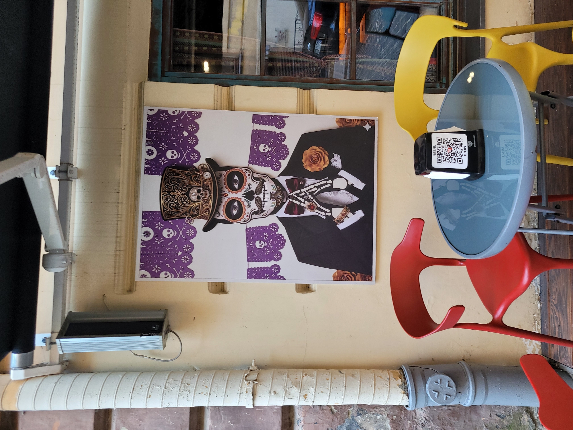 An outdoor cafe scene features a poster of a skeleton man in a top hat with intricate designs and skull motifs, wearing a suit and bowtie. He has skull face paint and skeleton hands. The poster is hung on a light yellow wall. In front of the poster, there is a small round glass-topped table with a gray metal frame. A black napkin holder with a QR code and "MENU" is placed on the table. Two chairs are positioned around the table: one is bright red and the other is bright yellow. To the left of the poster, a large, corrugated pipe runs vertically down the wall. The cafe is situated on a wooden floor. The overall ambiance suggests a festive or themed establishment.