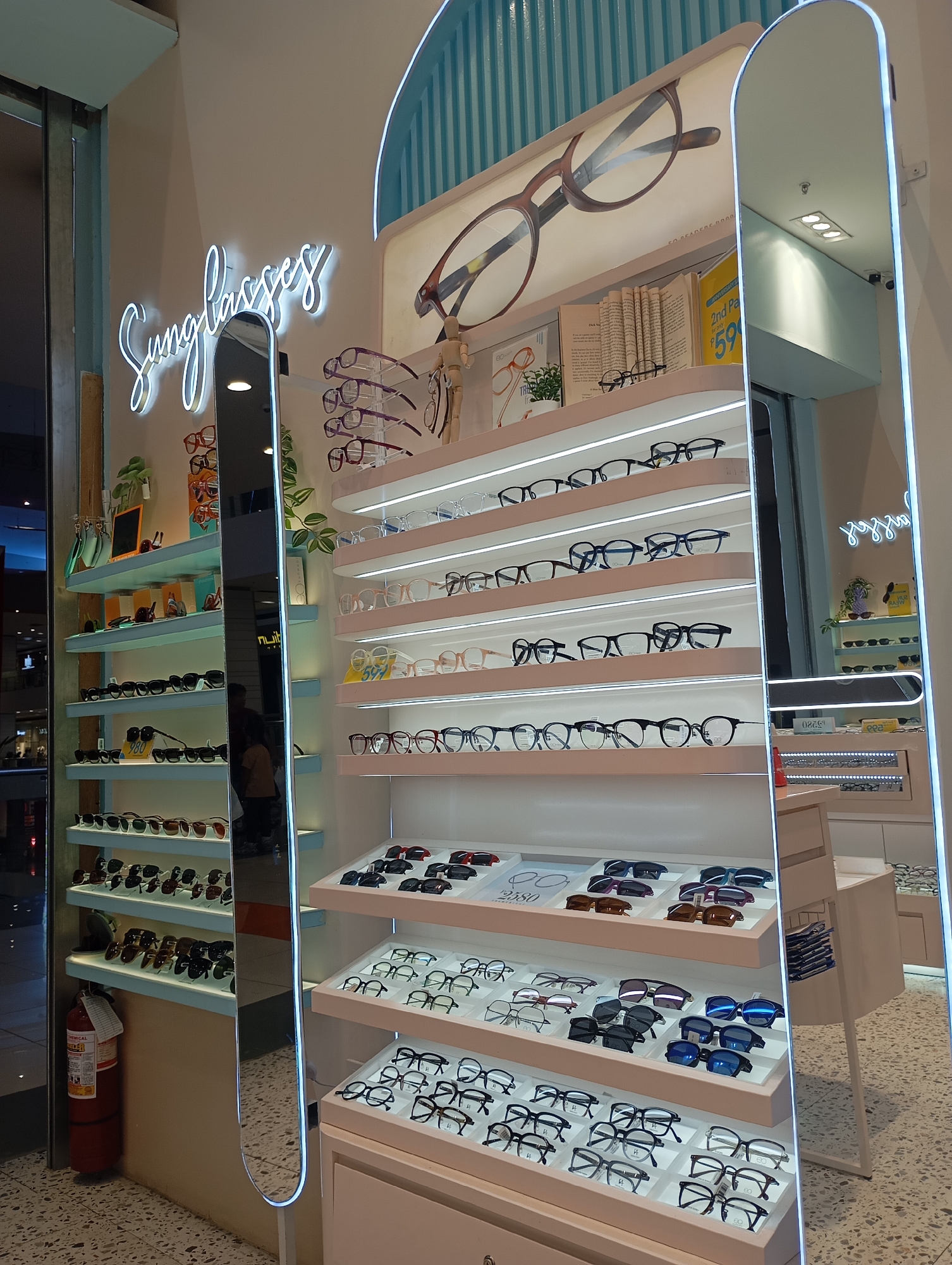 This image captures an interior view of a modern eyewear store, likely located within a mall in Davao City, Philippines. The scene is brightly lit by artificial lighting, including LED strips illuminating display shelves and a prominent neon sign.
The primary subjects are numerous pairs of eyeglasses and sunglasses, meticulously displayed on multi-tiered, light-colored shelves. On the left side, sunglasses are arranged on a series of shelves against a mirror, with a neon sign above them spelling "Sunglasses" in white cursive script. Below these shelves, a red fire extinguisher is visible on the floor.
The main display area on the right features multiple rows of eyeglasses, with each pair neatly placed on individual slots on illuminated shelves. Some shelves also hold decorative items like small potted plants, books, and a small wooden mannequin bust wearing glasses. A large, arch-shaped full-length mirror with glowing edges stands vertically on the right, reflecting parts of the store and a person wearing a dark top and light pants, possibly a customer or staff member. The floor has a light-colored, speckled terrazzo-like pattern.
Visible text includes the neon "Sunglasses" sign. On one of the book props, a price promotion reads "2nd Pa" and "only P59". Another small sign on the left display shelf shows "980", and a card on the bottom right shelf shows "2580", likely indicating prices. The brand "EQ" is visible on some glasses frames. The fire extinguisher is labeled "CHEMICAL EXTINGUISHER". The store appears to be clean and organized, designed for browsing eyewear. No specific event is taking place, but the setup suggests a retail shopping environment.