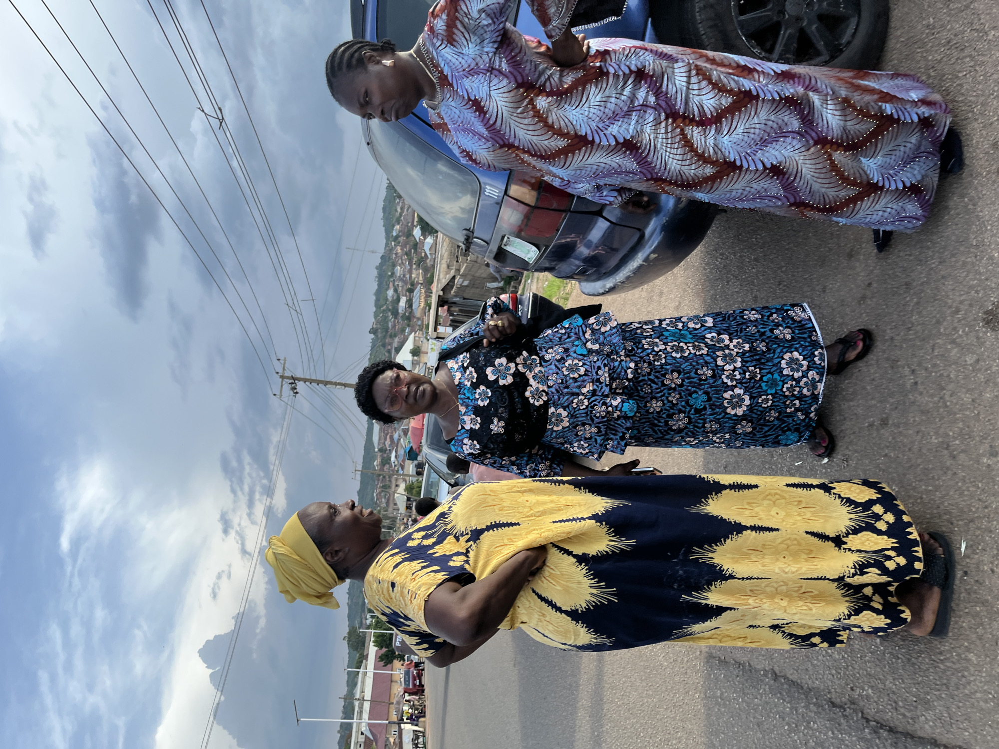 Three Nigerian women are standing on a paved street in Jos, Nigeria, conversing next to a dark blue vehicle. The woman on the left wears a yellow headscarf and a long yellow and navy blue patterned dress, with her left hand on her hip. The woman in the center wears a black headwrap and a blue floral print dress. The woman on the right wears a long, multi-colored patterned dress in shades of purple, blue, and red, with her right hand on her hip. The background shows a hazy, overcast sky, utility poles with wires, and a residential area with buildings and hills. The street appears to be in a moderately populated urban or semi-urban area.
