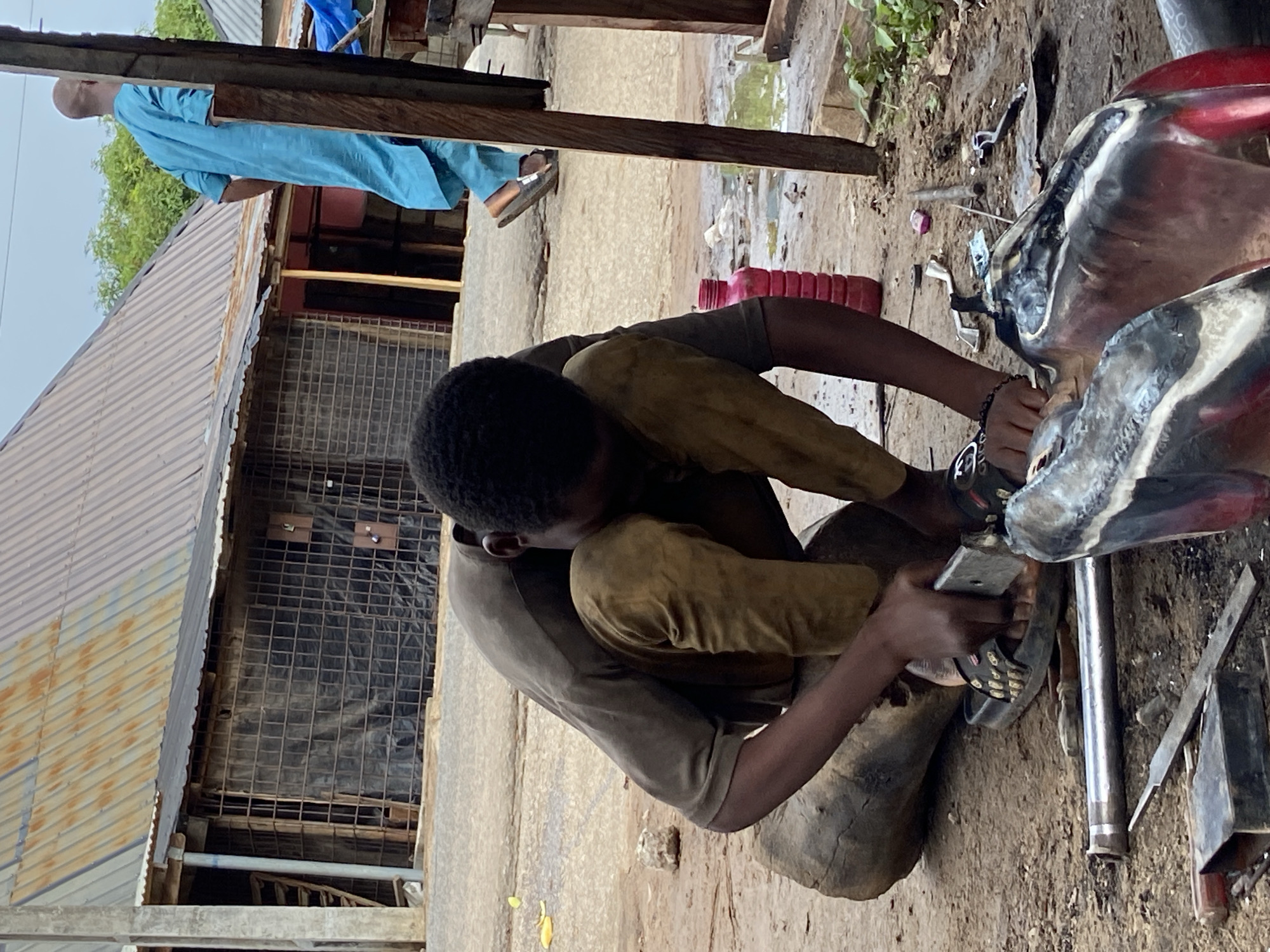 A young man is working on a motorcycle, likely repairing or modifying it. He is squatting on the ground next to the motorcycle, which is in a partially disassembled state, with tools and parts scattered around. The scene is in an outdoor setting, likely a repair shop or workspace in Mararaba, Nigeria, based on the visual context. Another man is seen standing in the background. The surroundings are a mix of urban and possibly somewhat rural elements, with buildings of varying construction quality nearby.