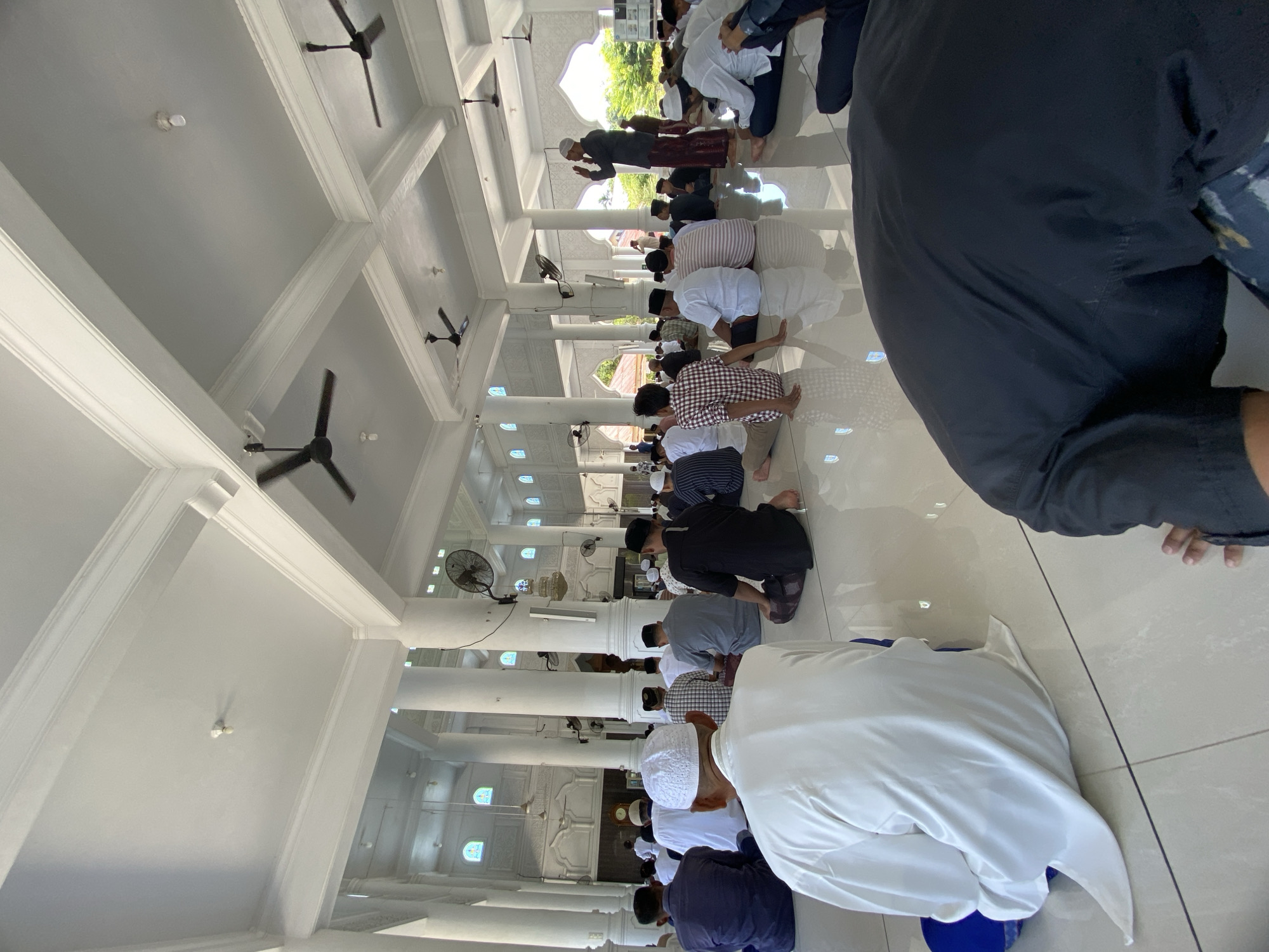Several people are kneeling on a light-colored tile floor in what appears to be a mosque. The building has white pillars and a white ceiling with dark ceiling fans. In the background, there are stained-glass windows. One person is standing at the end of the line, facing the other people. This location is in Lhokseumawe, Indonesia.