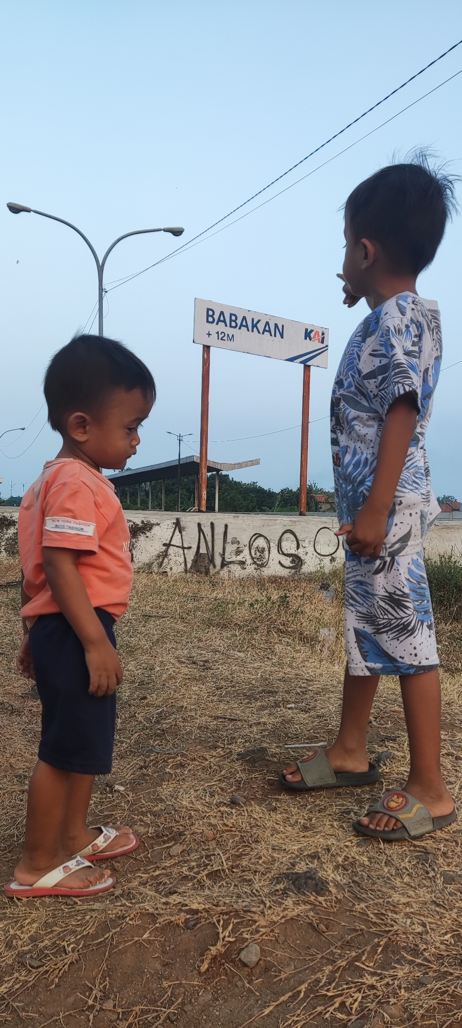 Two young boys stand on dry, yellowed grass in front of a concrete wall with graffiti. The boy on the left wears an orange t-shirt and dark blue shorts with white flip-flops. He looks down and to his left. The boy on the right wears a patterned t-shirt and shorts with grey sandals. He looks up and to his right, with his right arm extended slightly. Behind them, a sign reads "BABAKAN + 12M KAI," indicating a location in Babakan, Pakusamben, Indonesia. The background shows a street lamp and some trees.