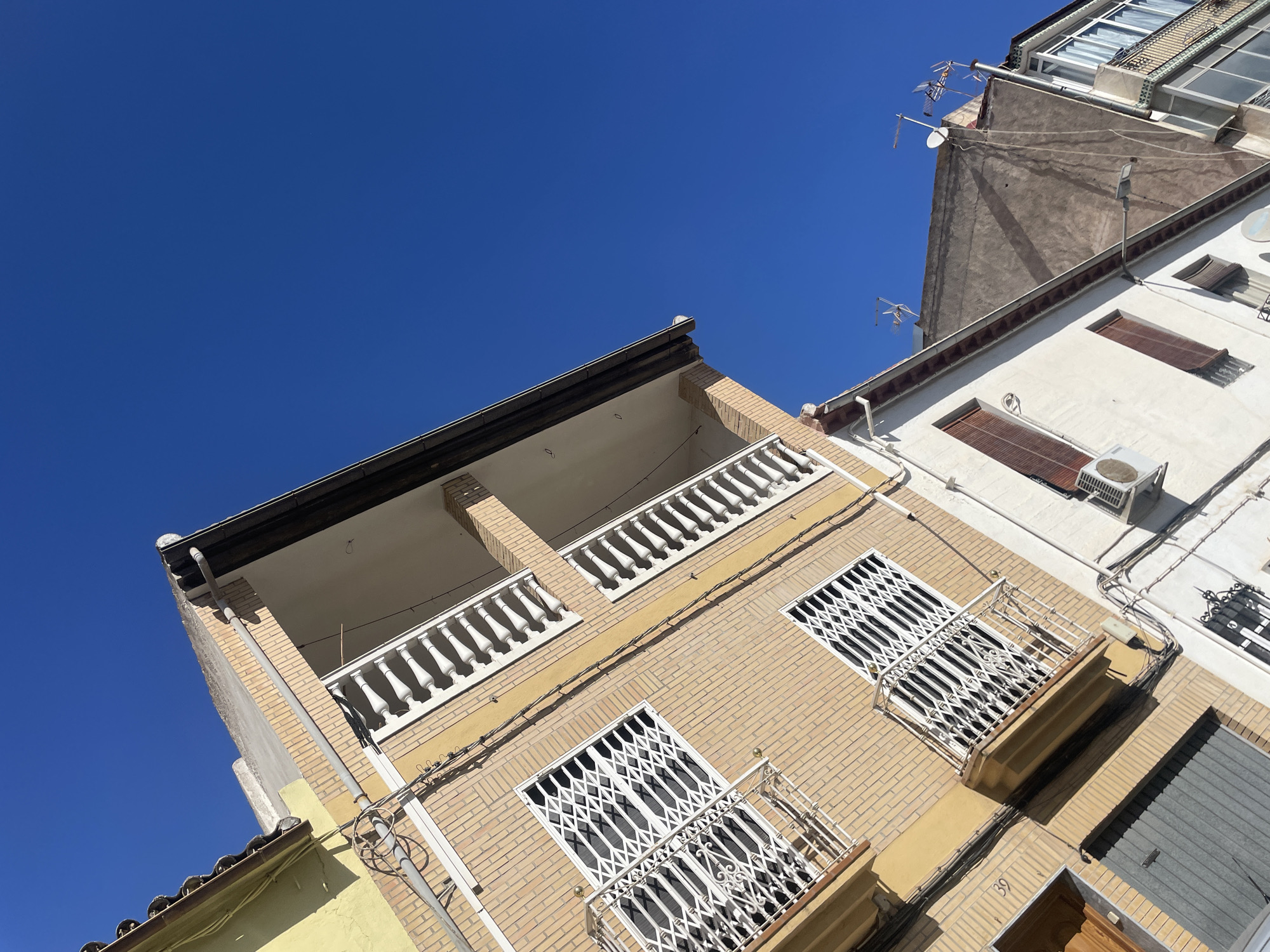 The image shows a street scene in Atarfe, Spain, characterized by an upward angle that emphasizes the facades of residential buildings against a clear blue sky. The main building in the foreground is constructed with light brown bricks, featuring multiple balconies with white balustrades. The windows on this building are protected by white metal security grates. To the left of this building, a portion of a yellow-painted facade is visible, along with some electrical wiring and a round metal fixture. The adjacent building to the right is painted white and has brown-shuttered windows and an outdoor air conditioning unit. Further in the background, another building with a grey, textured facade is seen, topped with several television antennas and satellite dishes. The scene depicts typical urban architecture under bright sunlight.