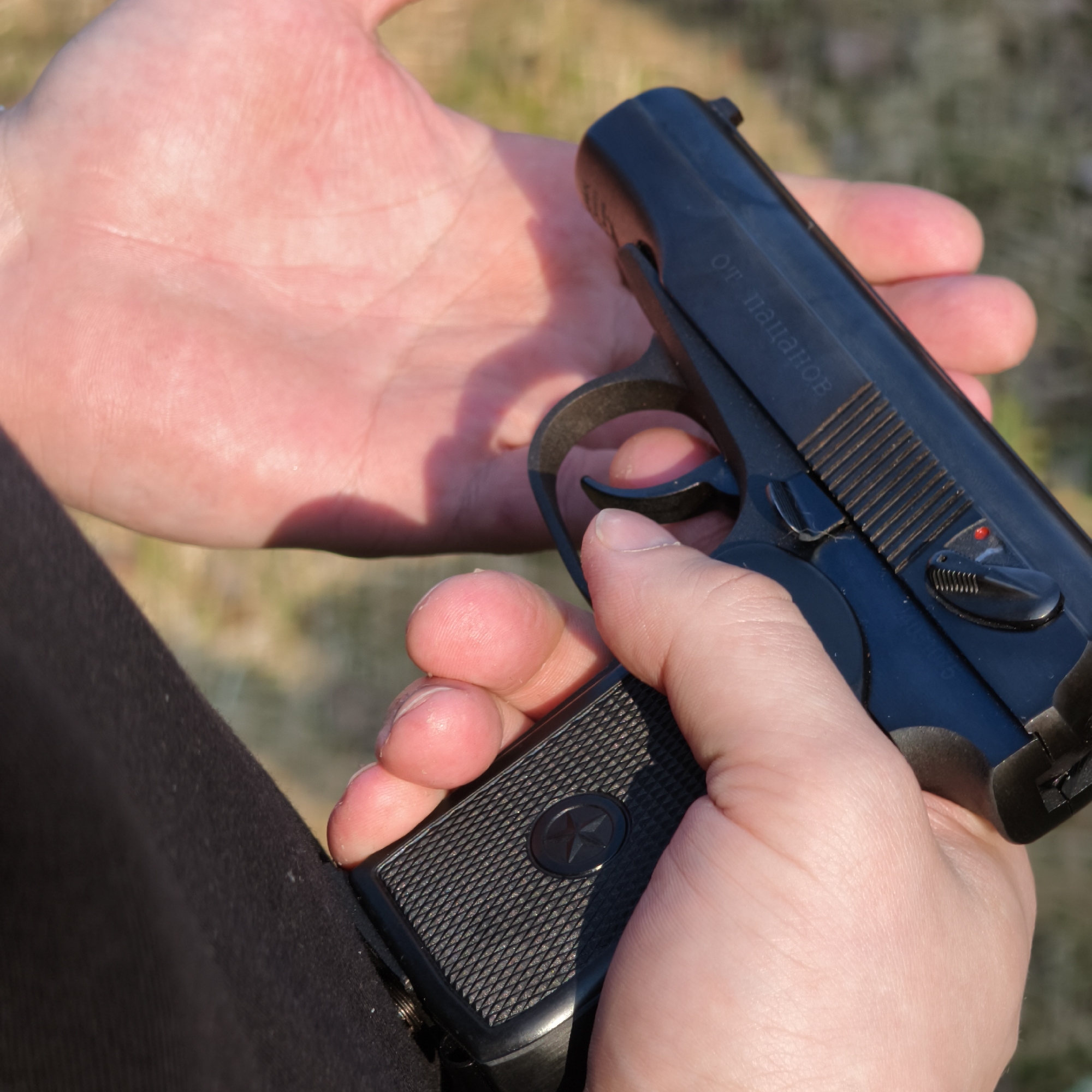 The image depicts a close-up view of a black semi-automatic pistol being held by two hands, with an out-of-focus outdoor background, located in the city of Парэчча, Belarus. The right hand grips the pistol's checkered black handle, which features a central five-pointed star emblem. The index finger of the right hand is positioned around the trigger. The pistol's slide displays Cyrillic text, "ОТ ПАЦАНОВ" (OT PATSANOV - "FROM THE BOYS"), and a visible red dot indicates the safety lever is in the 'fire' position. The left hand is partially visible in the upper background, cupped. A dark textile material is visible on the lower left, adjacent to the pistol's grip.