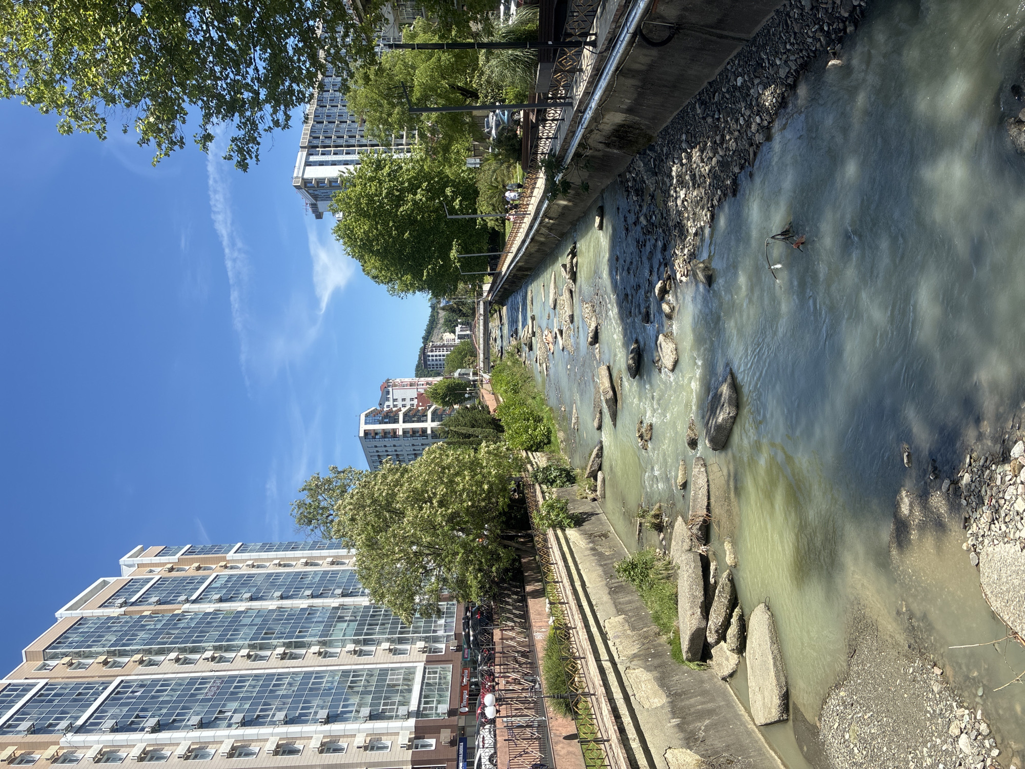 This image captures a sunlit urban scene in Sochi, Russia, featuring a river flowing between modern city developments. The perspective looks down the length of the river, which appears shallow with numerous rocks and stones visible in its murky, green-grey water. Concrete embankments line both sides of the waterway.
On the left side of the river, a paved pedestrian promenade runs parallel to the bank, protected by a decorative metal fence with geometric patterns. Several individuals are visible on this path, appearing to walk or stand. Behind the promenade, a dense line of lush green trees, some with white blossoms, provides shade. Dominating the background on this side are tall, contemporary buildings with extensive glass windows and light-colored facades, many featuring visible air conditioning units.
The right side of the river also has a concrete embankment, beyond which more buildings and greenery can be seen in the distance, though less distinct due to perspective and distance. The sky above is a clear, bright blue with a few thin, wispy clouds, indicating fair weather. The strong sunlight suggests it is midday or early afternoon. The overall scene conveys a pleasant, lively atmosphere in a modern city during warm weather. No legible text is visible.