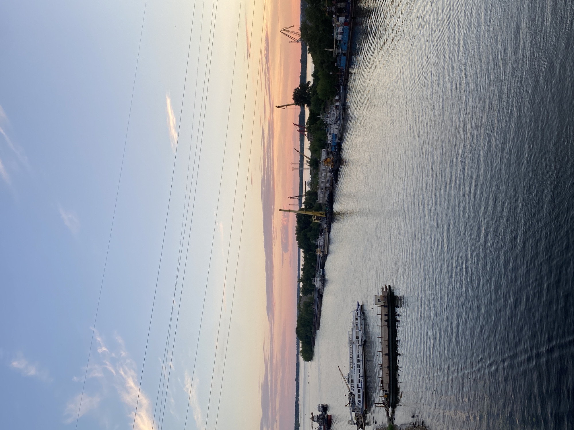 This is a vertical, high-angle shot of a river with a city skyline and a cloudy sunset. The sky is a pale blue with streaks of pink and orange from the setting sun, and several thin power lines stretch across the top of the frame. On the left side of the river is a city with tall buildings and trees. Several boats and ships are docked along the shore, and construction cranes are visible throughout the city. The water in the river is dark and choppy, with reflections of the sky and buildings. The scene appears to be from the early evening, possibly during the summer, given the presence of green trees and the warm sunset colors. The overall mood is calm and peaceful. There is no visible text in the image.